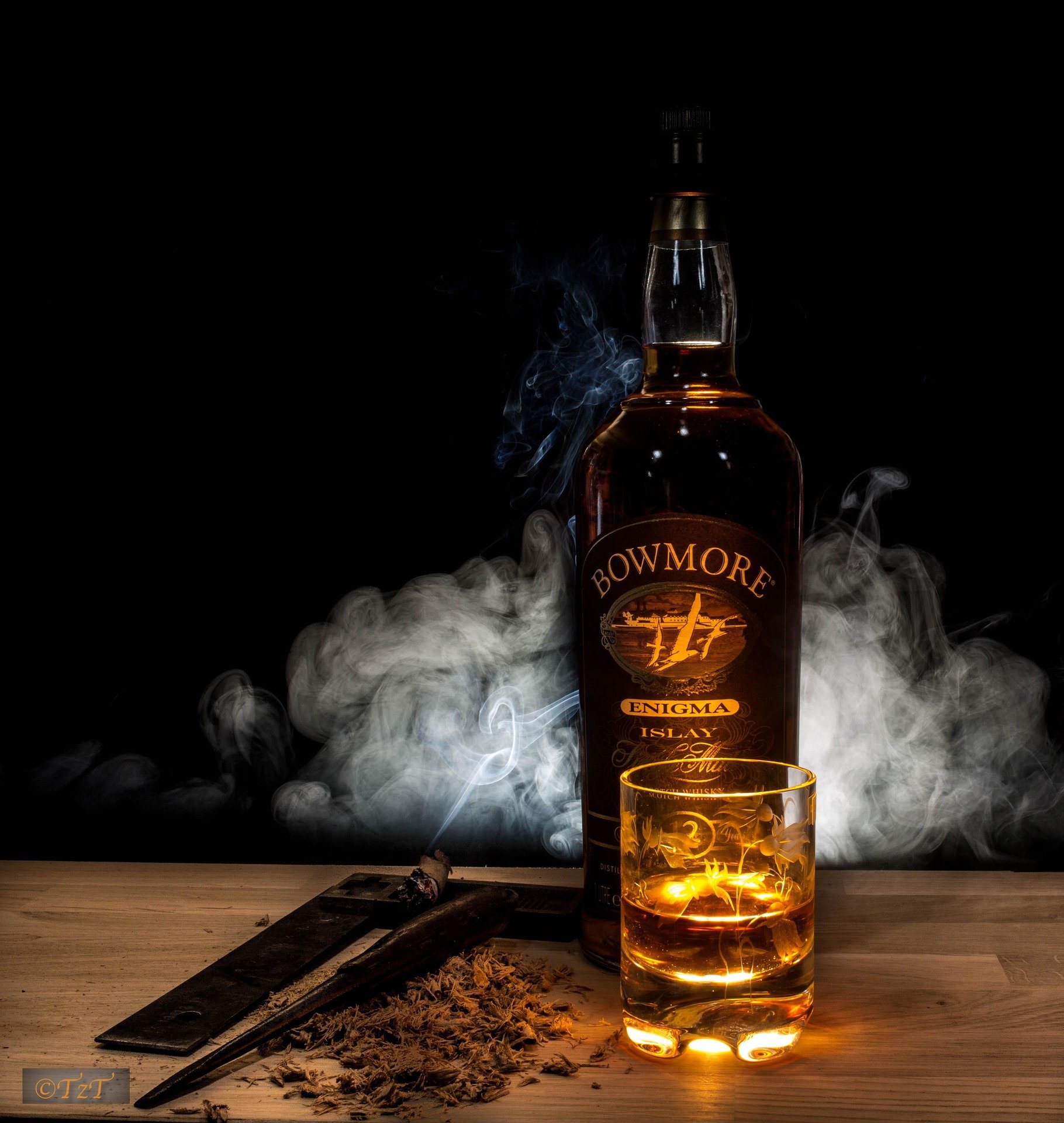A bottle of Bowmore Enigma Islay Scotch whisky, a glass filled with whisky, a lit cigar resting on a wooden surface with ash and a cigar cutter, and smoke swirling around on a dark background.
