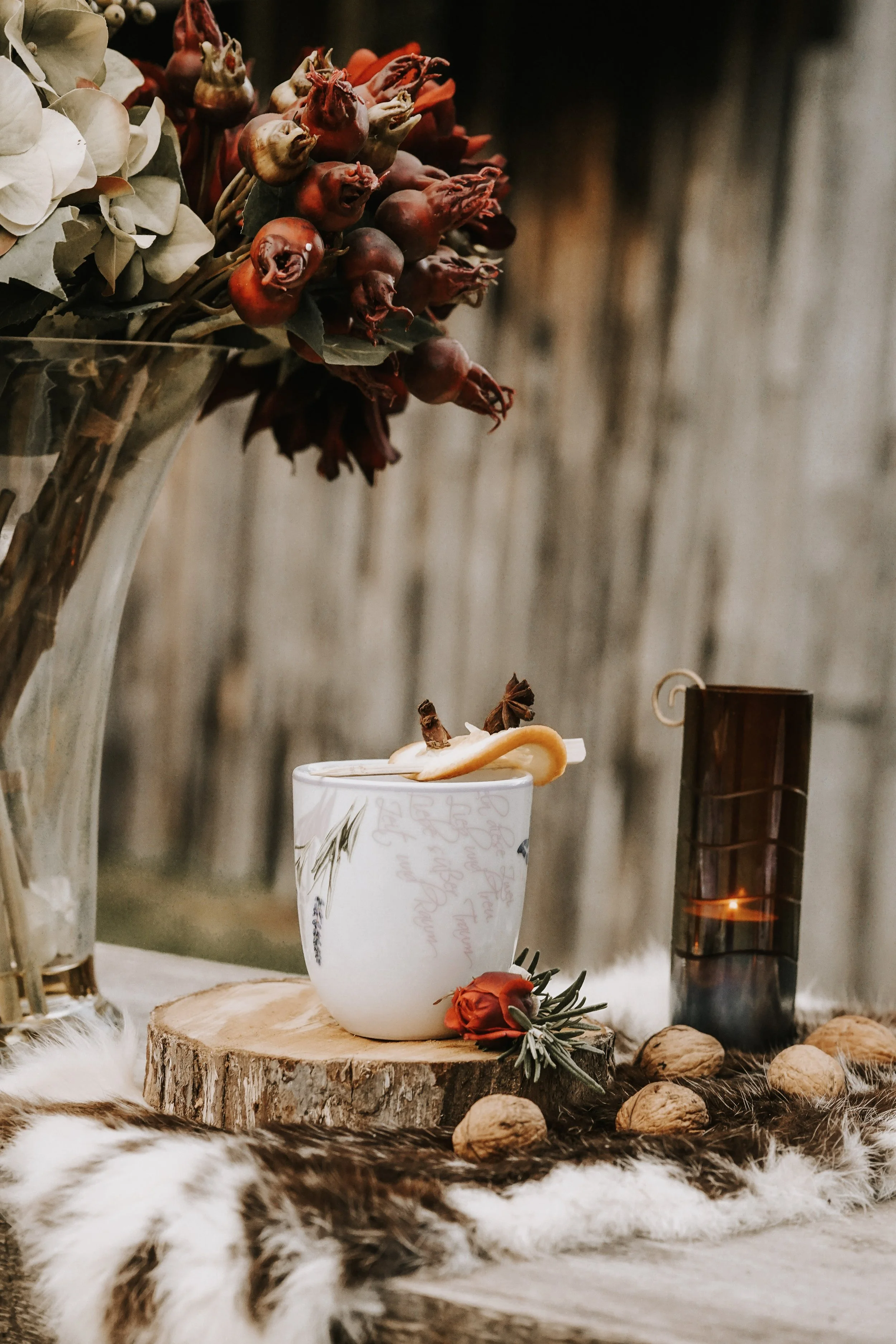 A decorative still life scene with a white cup, a yellow lemon peel, and star anise on a wooden slab, surrounded by walnuts, a small red rose, and pine needle sprigs, with a tall candle holder, dried flowers, and a fur rug in the background.