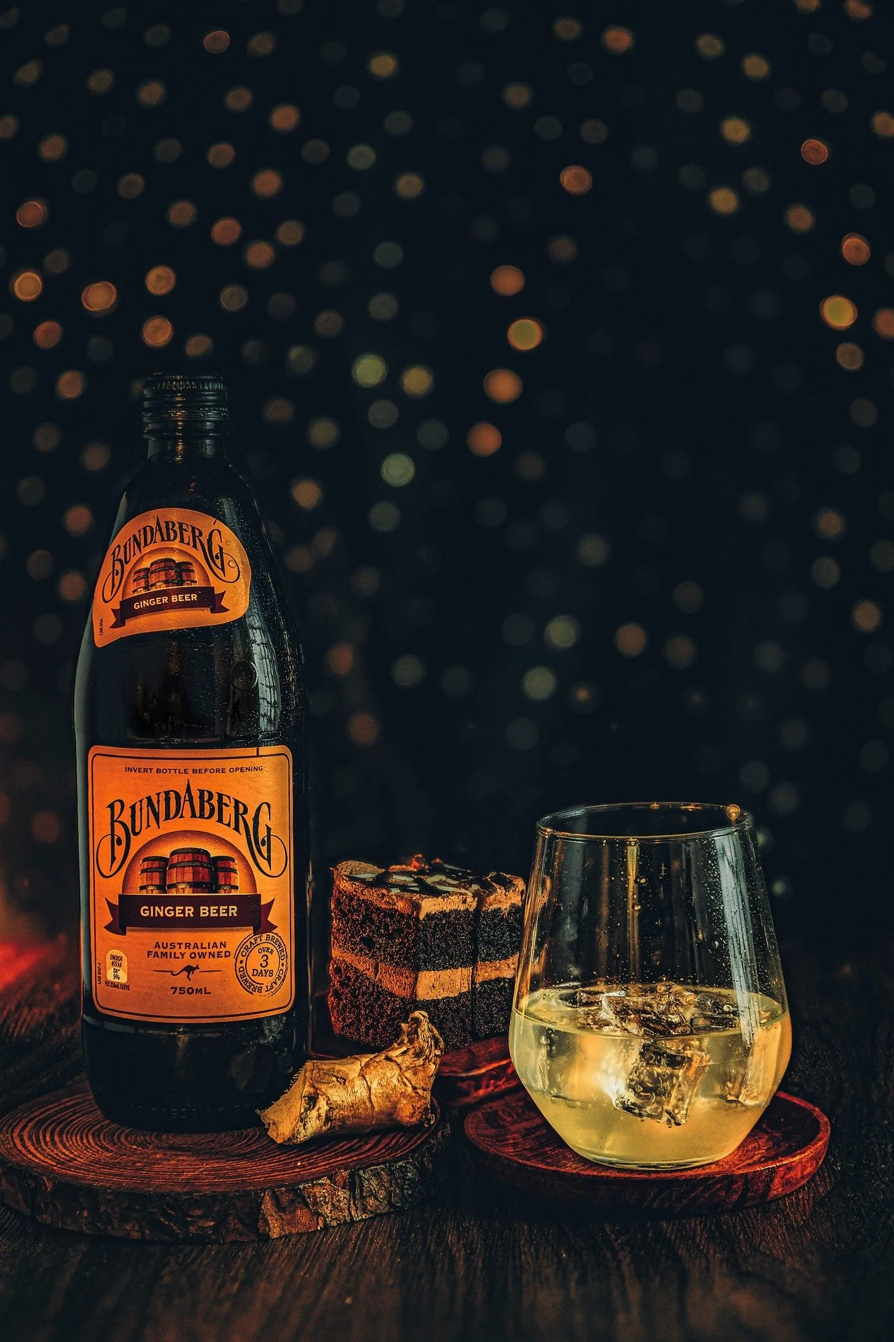A bottle of Bundaberg ginger beer, a glass of ginger beer with ice, a piece of dark layered cake, a piece of ginger root, and a chocolate cake with multiple layers, all arranged on wooden coasters against a dark background with bokeh lights.