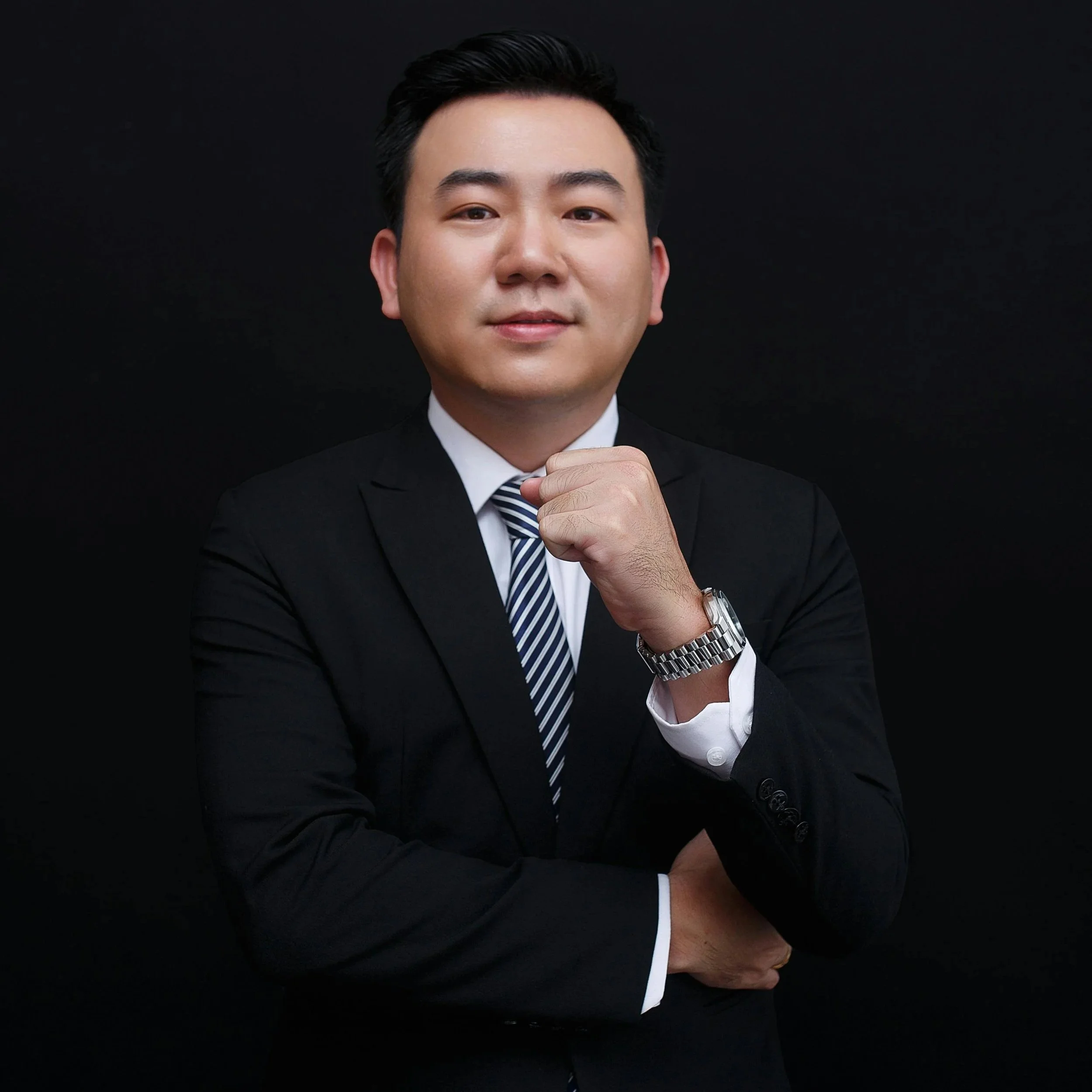 Professional man in a suit with a striped tie, wearing a watch, posing with his hand near his chin against a black background.