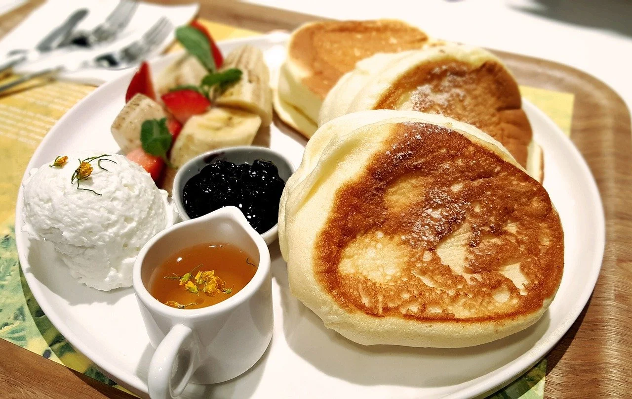 A plate with two golden-brown pancakes, a side of whipped cream topped with small flowers, fresh sliced bananas and strawberries, a small bowl of dark berry jam, and a small pitcher of syrup with yellow flowers floating inside.