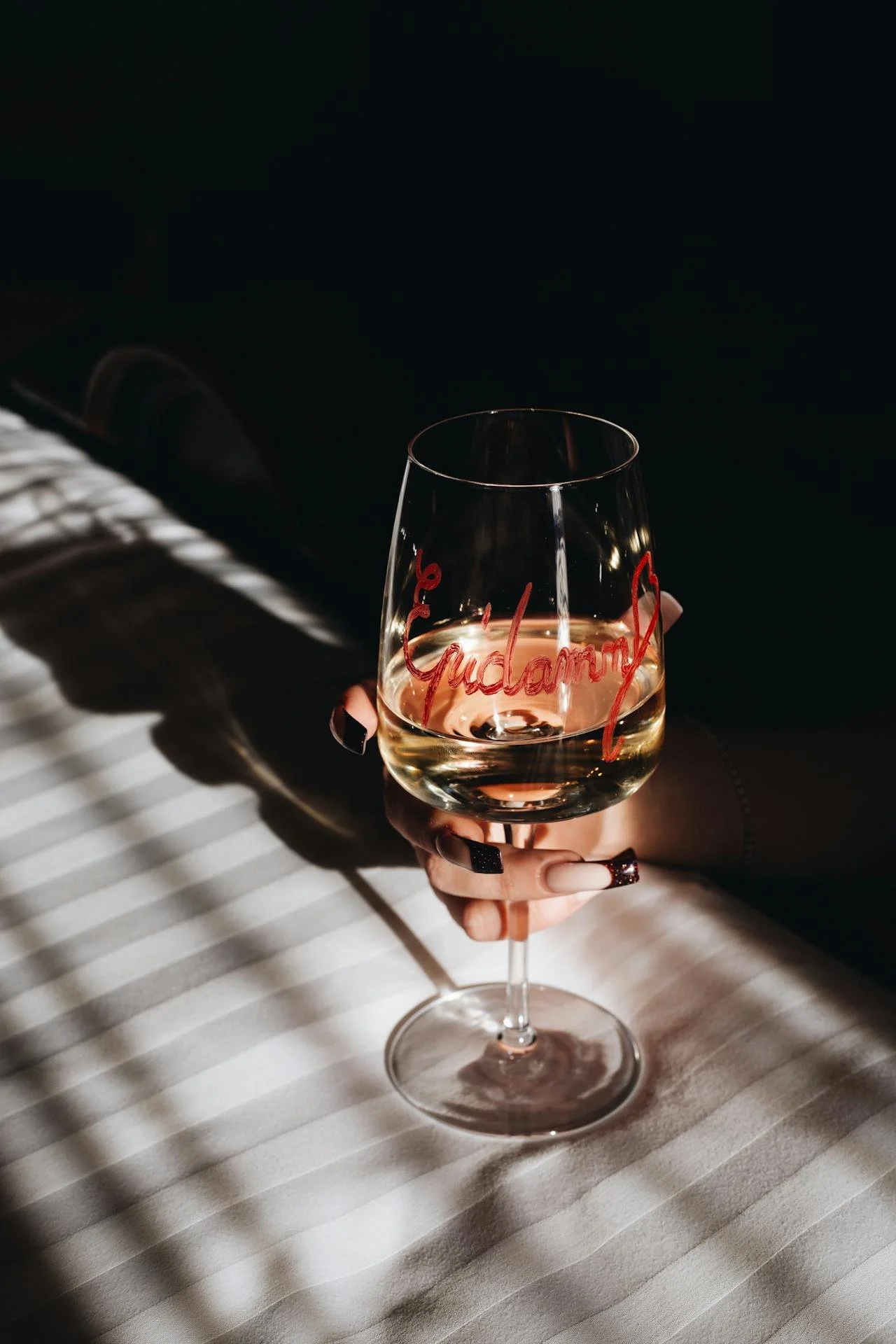 A hand holding a glass of white wine with the word 'Guilty' written on it in red. The glass casts a shadow on a striped tablecloth, with sunlight creating patterns on the surface.