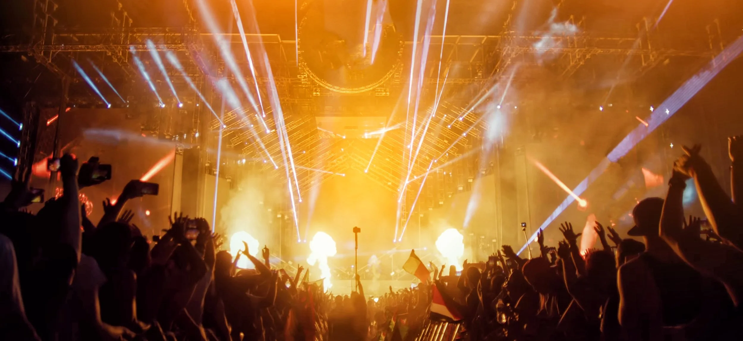 Crowd of people at a concert or music festival with bright stage lights, pyrotechnics, and laser beams illuminating the scene.