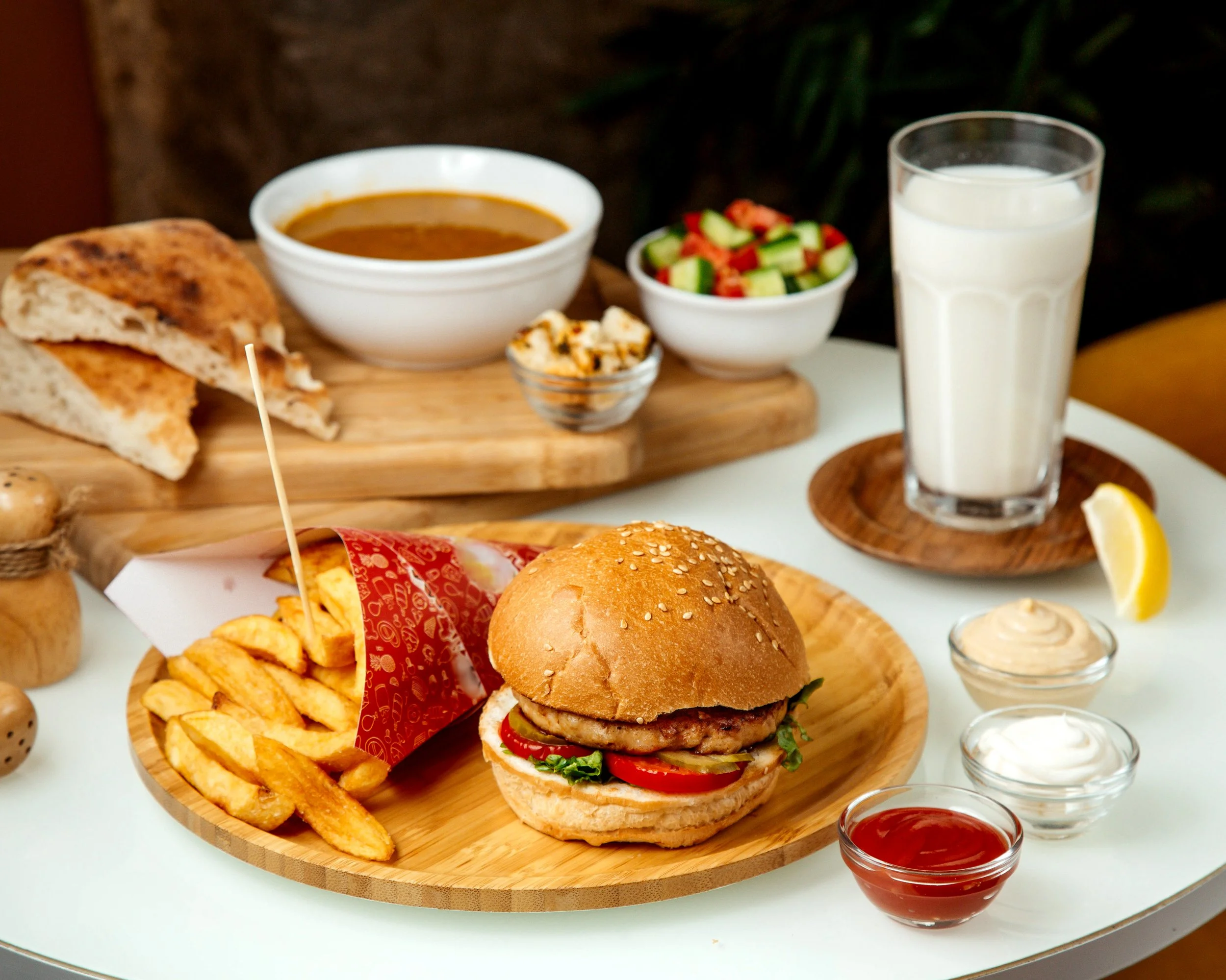 A burger with fries, condiments, and a lemon wedge on a wooden plate, with a side salad, soup, and a glass of milk in the background.