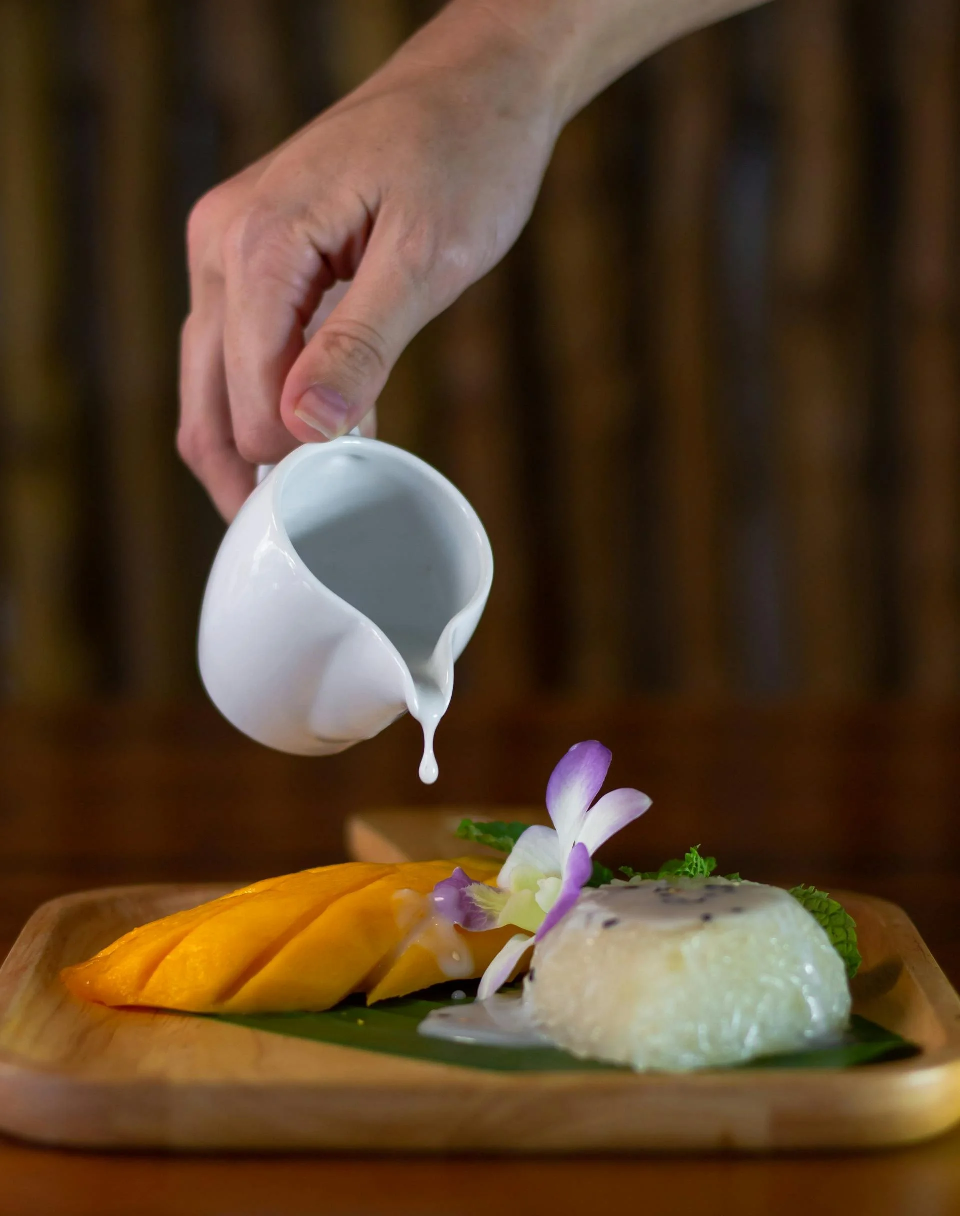 Person pouring a white sauce or cream over a mango and a dessert garnished with an orchid flower on a wooden tray.
