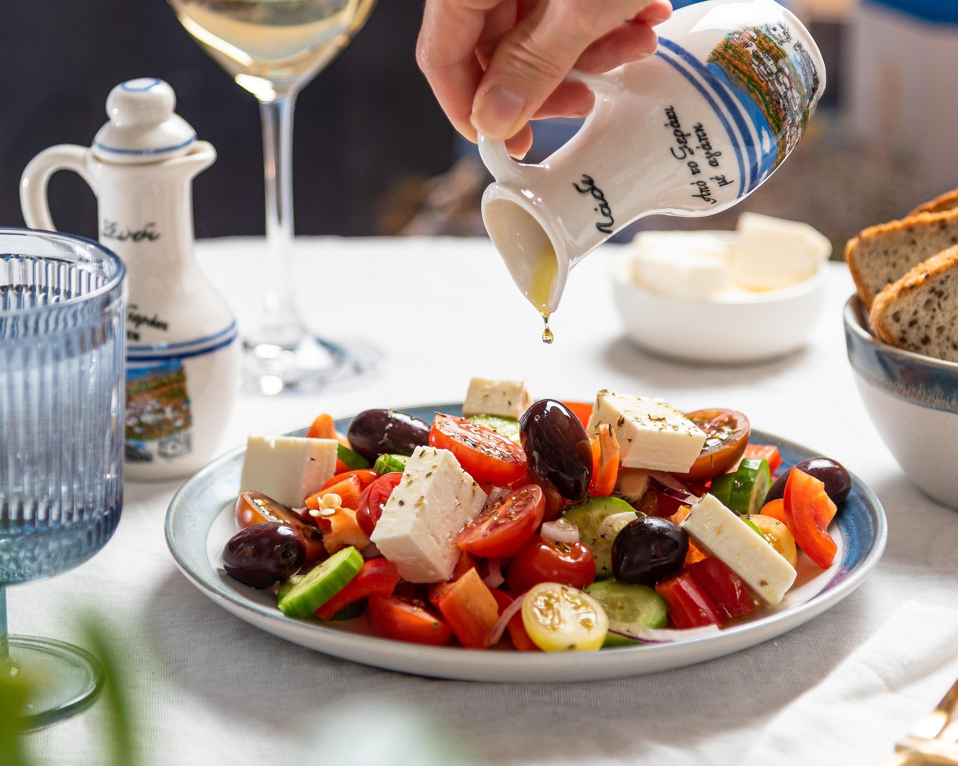 A person pouring olive oil onto a Greek salad with tomatoes, cucumbers, olives, and feta cheese, on a white table with wine, bread, and condiments in the background.