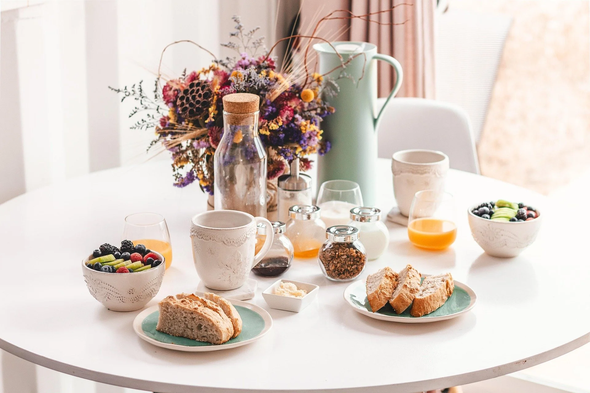 A breakfast table with bowls of fresh fruit, slices of bread, coffee mugs, glasses of orange juice, and various jars of preserves and spreads, decorated with a large bouquet of colorful dried flowers in the background.