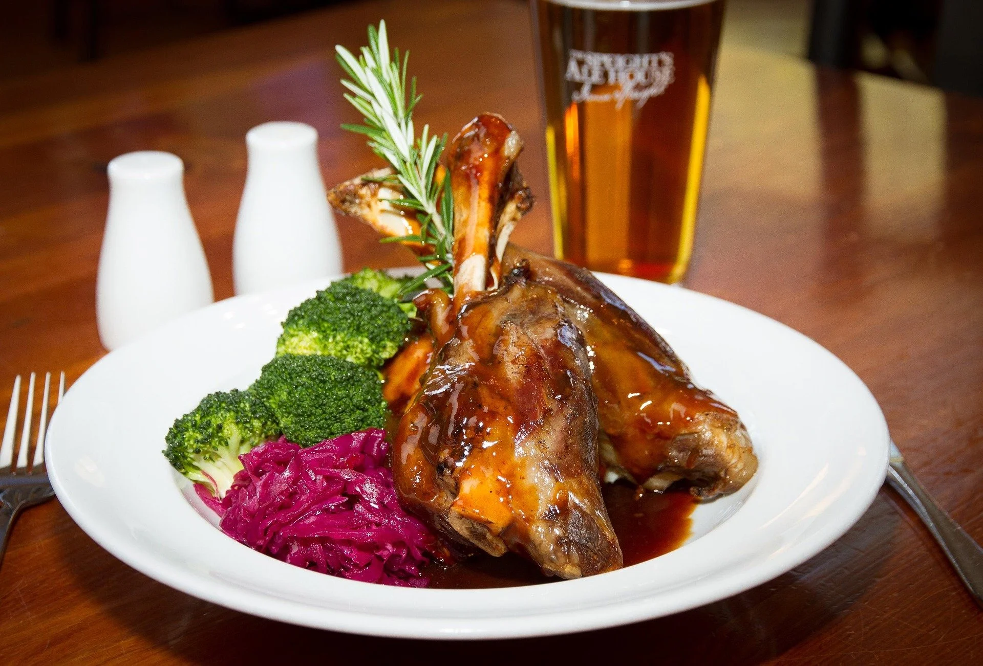 A plate with two glazed lamb shanks garnished with a sprig of rosemary, served with steamed broccoli and pickled red cabbage. In the background, there is a glass of beer and salt and pepper shakers on a wooden table.