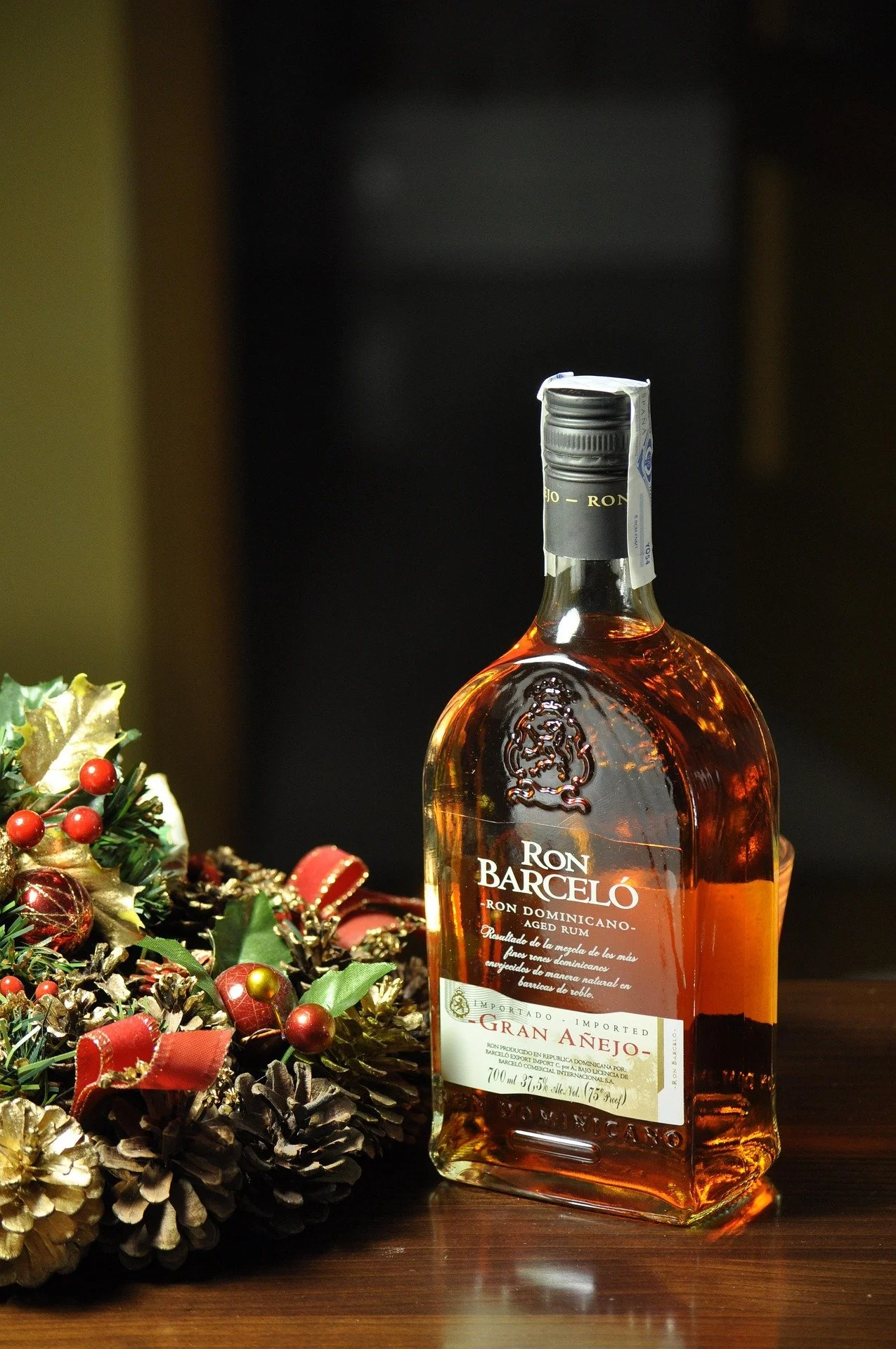 A bottle of Ron Barceló Gran Añejo rum placed on a wooden surface next to a Christmas wreath with red and gold decorations, with a dark background.