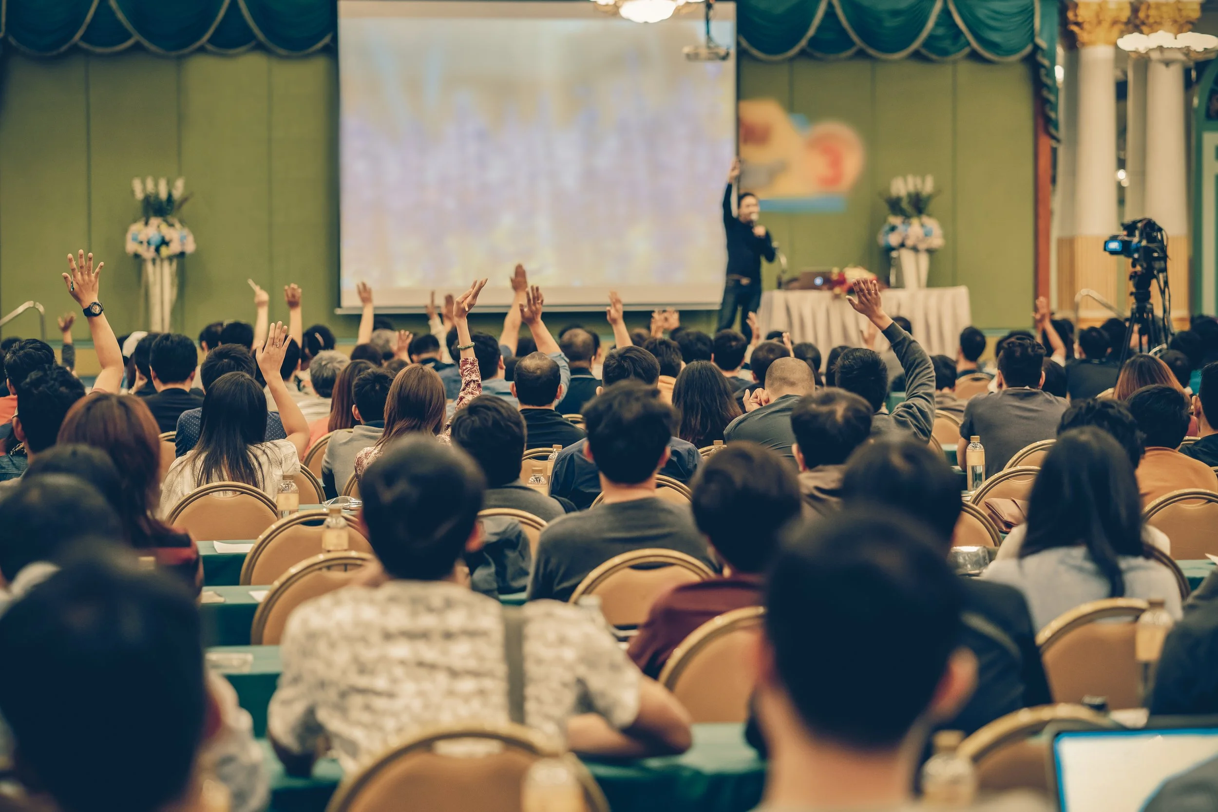 A large conference room filled with people attending a presentation. Many attendees have their hands raised while a speaker stands at the front of the room, near a large screen and a table with flowers, addressing the audience.