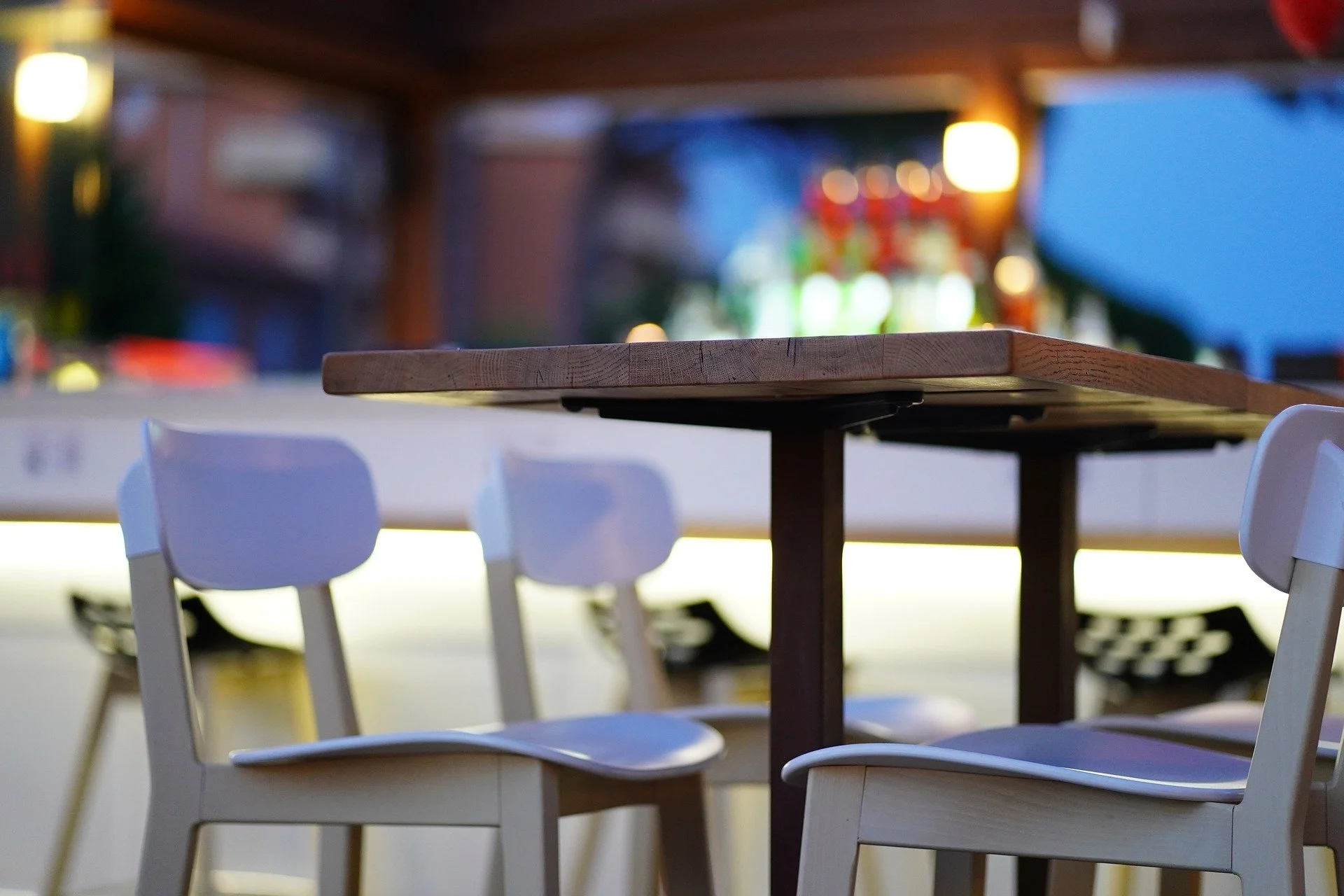 Empty outdoor dining area with white chairs and a wooden table at dusk, with blurred colorful lights in the background.