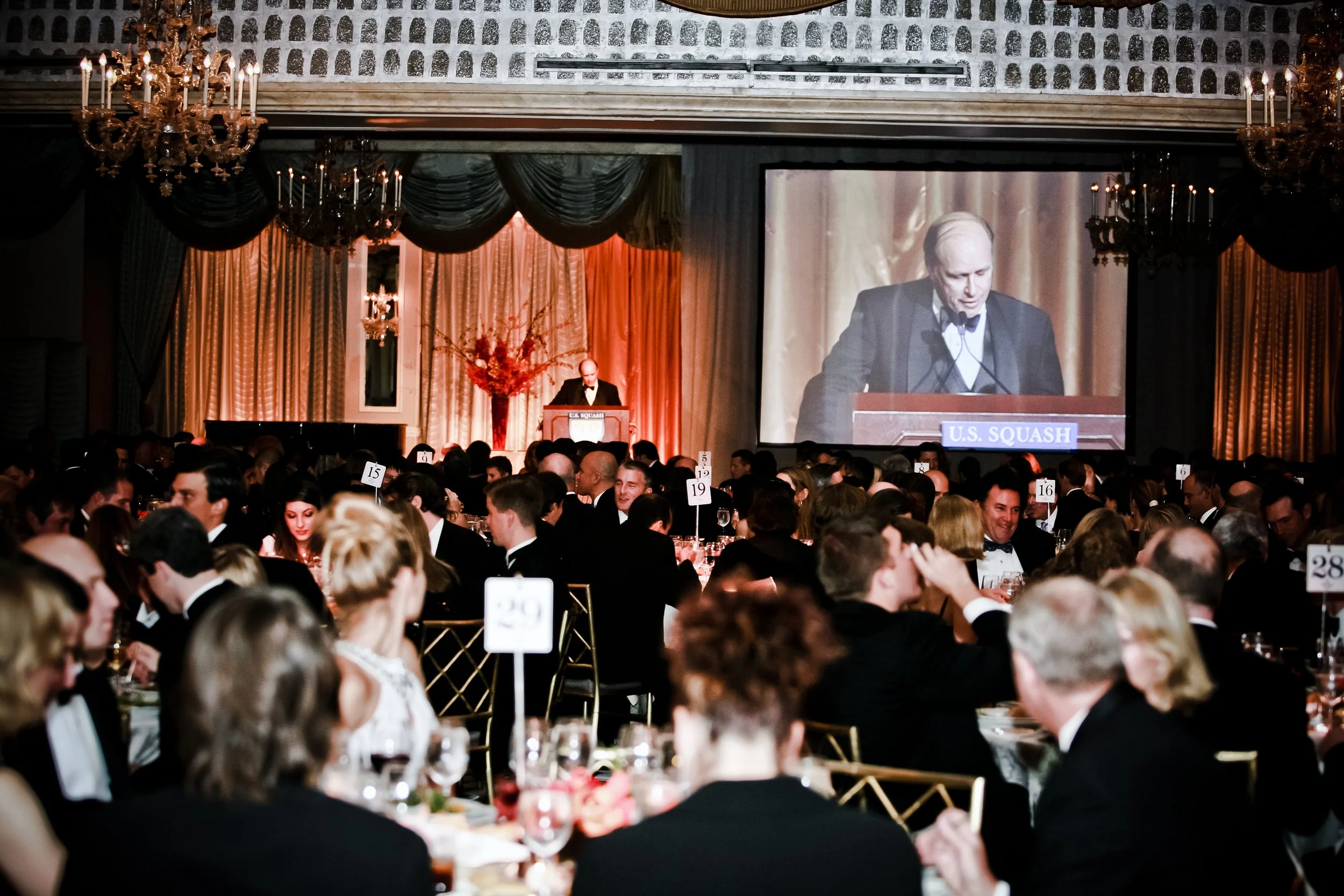 A large formal banquet hall filled with people dressed in tuxedos and evening gowns, attending a fundraising event for U.S. Squash. A man is speaking at a podium with a large screen behind him displaying his image.