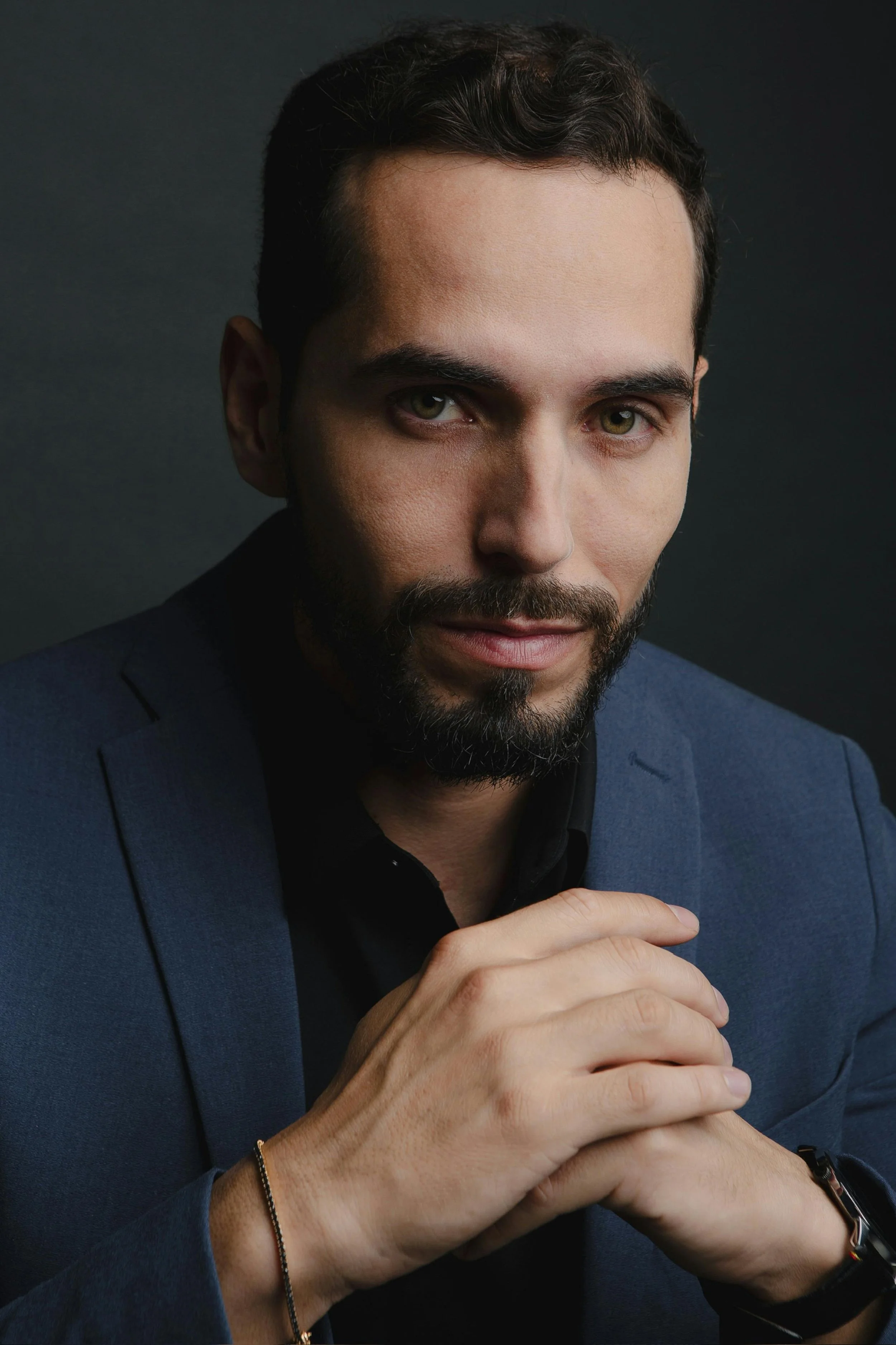 A man with dark hair and a beard wearing a dark blue blazer and black shirt, resting his hands together with a slight smile, against a dark background.