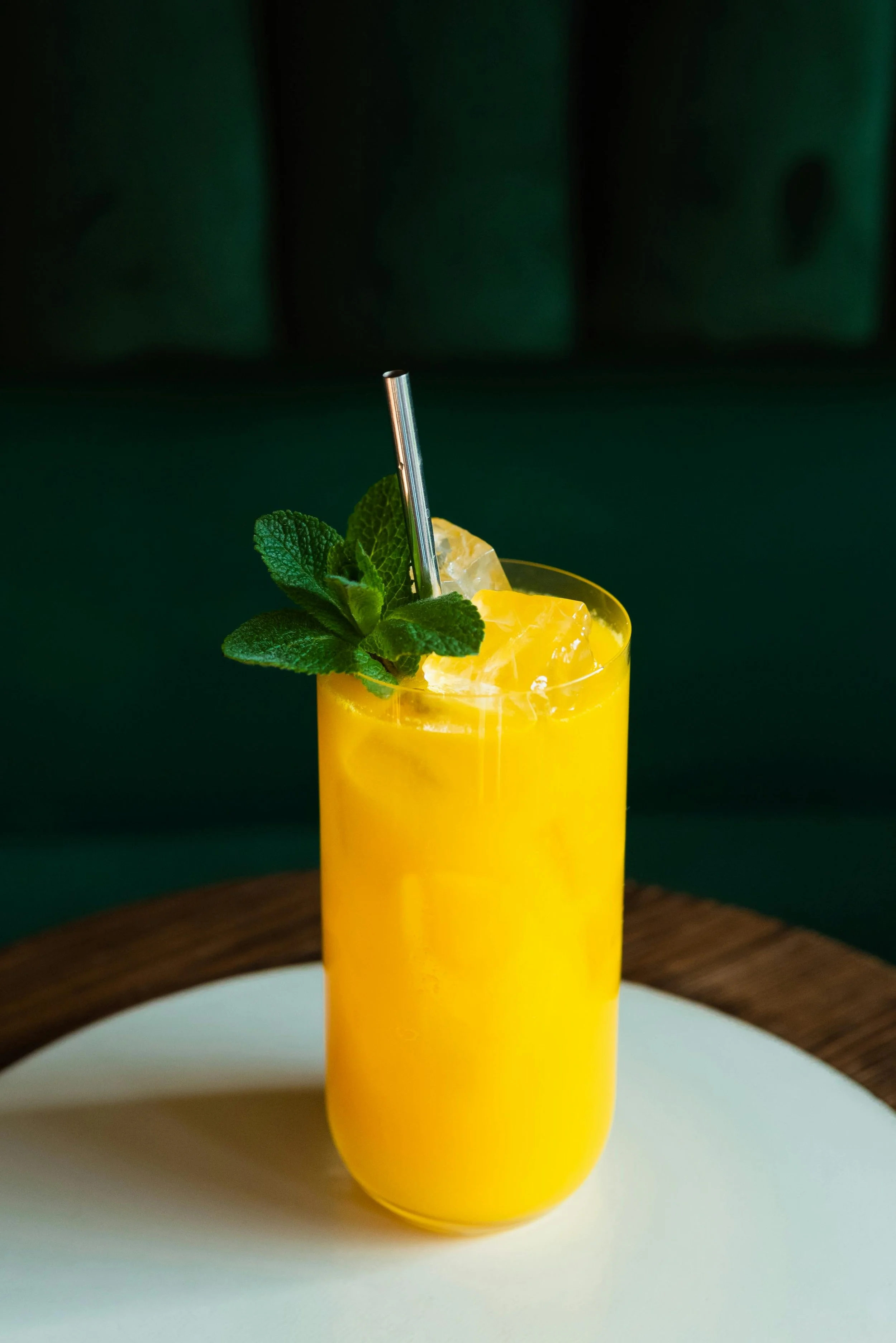 A tall glass of yellow mango juice garnished with fresh mint leaves, a slice of lemon, and ice cubes, with a metal straw inside the glass, on a white and wood table surface.