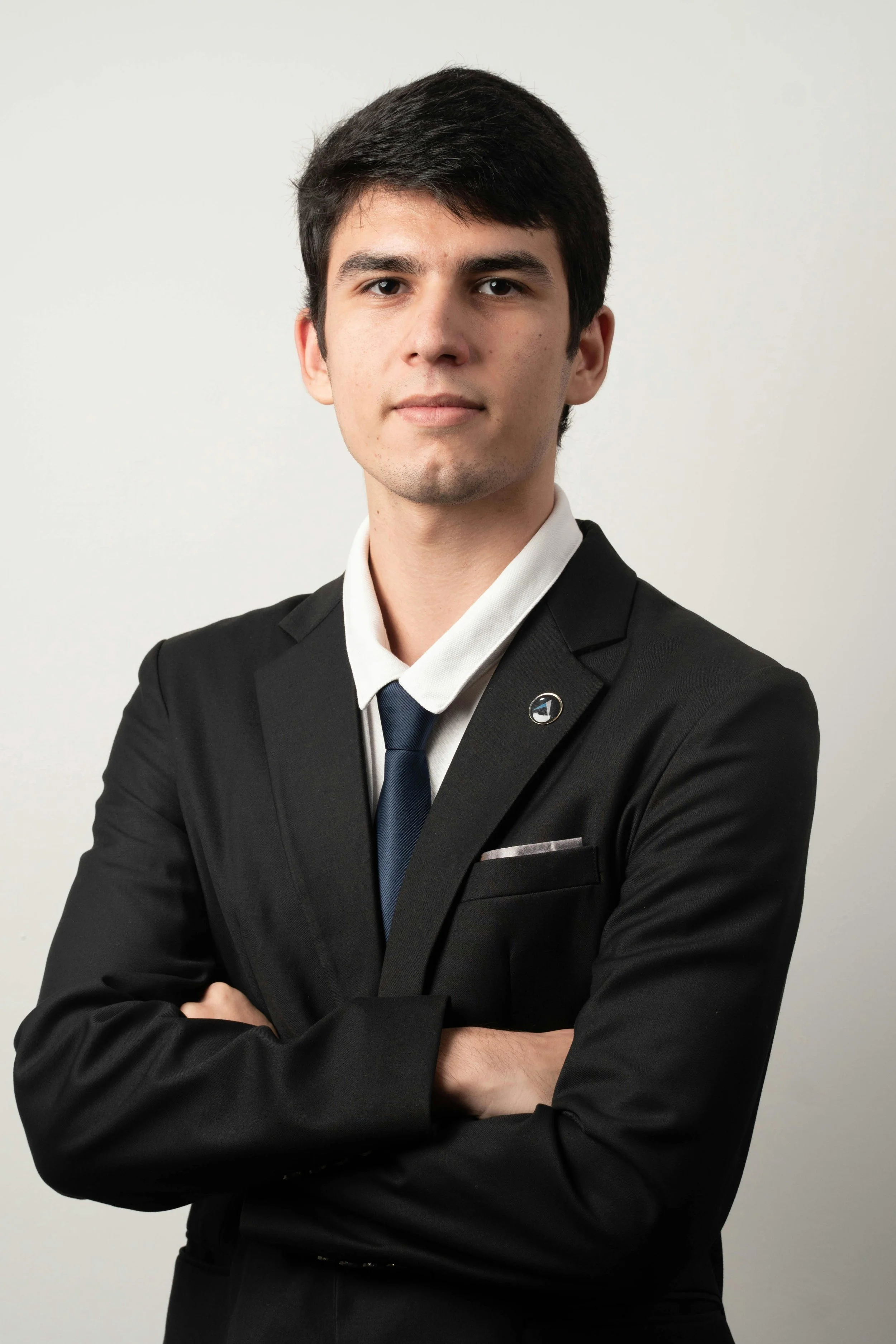 A young man with dark hair in a business suit, white shirt, and dark tie, standing with arms crossed against a plain light background.
