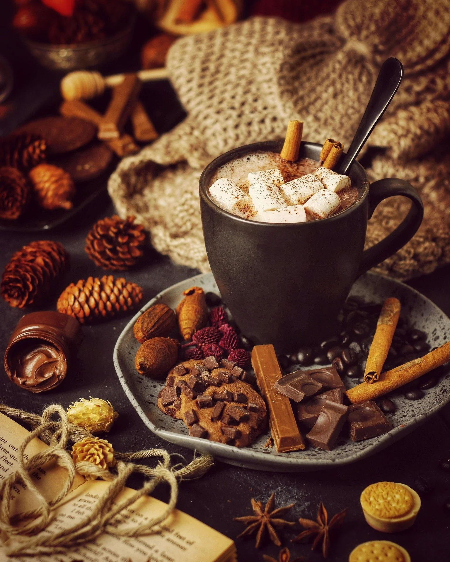 A hot chocolate in a black mug topped with marshmallows and cinnamon sticks, surrounded by assorted chocolates, cookies, nuts, and spices on a dark surface with a beige knitted cloth nearby.