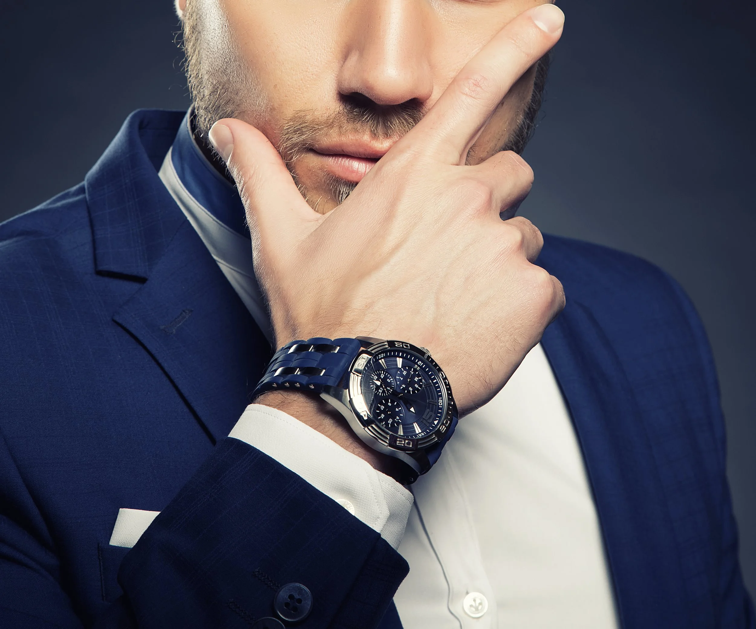 Close-up of a man in a blue suit with a white shirt, wearing a large black wristwatch, with his hand near his face and showing a thoughtful expression.