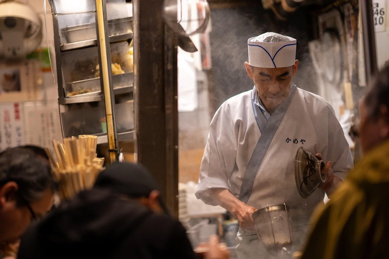 A Japanese chef wearing a traditional white uniform and hat preparing food at a busy restaurant kitchen, with steam rising around him and customers seated in front.