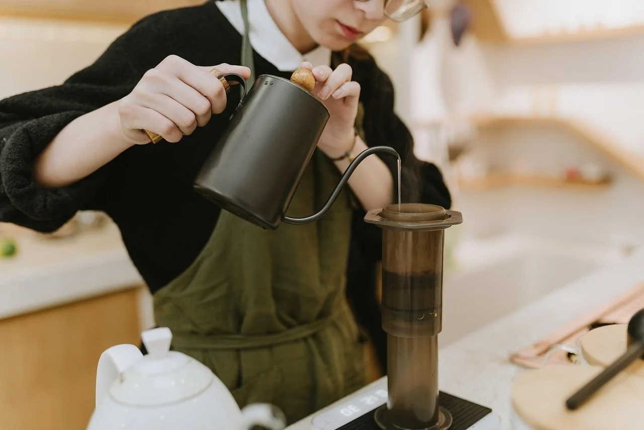 Person pouring hot water from a black kettle into a pour-over coffee maker on a kitchen counter.