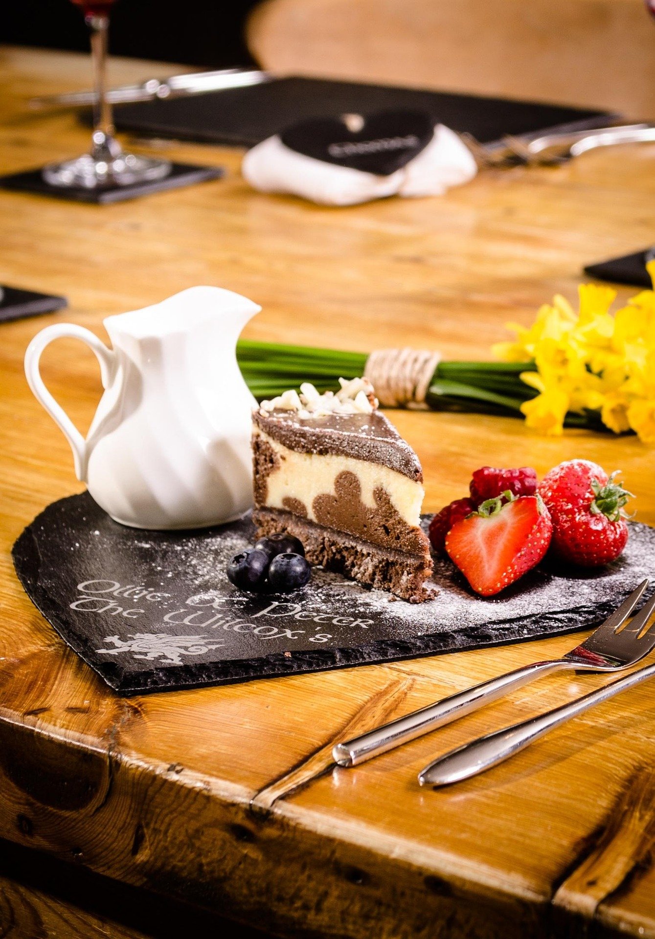 A dessert plate with a slice of cheesecake topped with chocolate, fresh strawberries, blueberries, and blackberries, served with a small white pitcher, on a black slate board with white writing, on a wooden table, accompanied by a fork and knife, wit