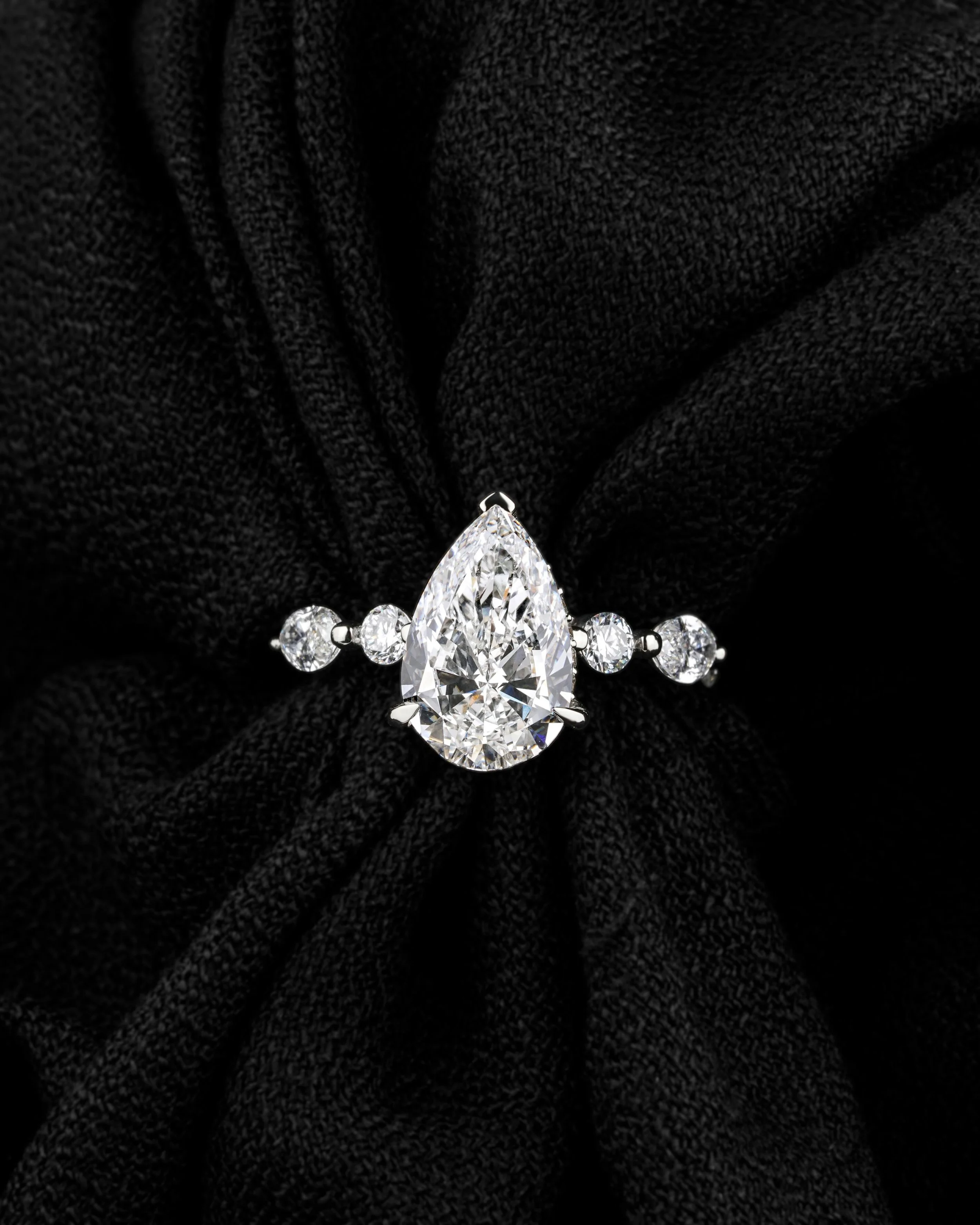 A diamond ring with a pear-shaped center stone and small round side stones on a black fabric background.