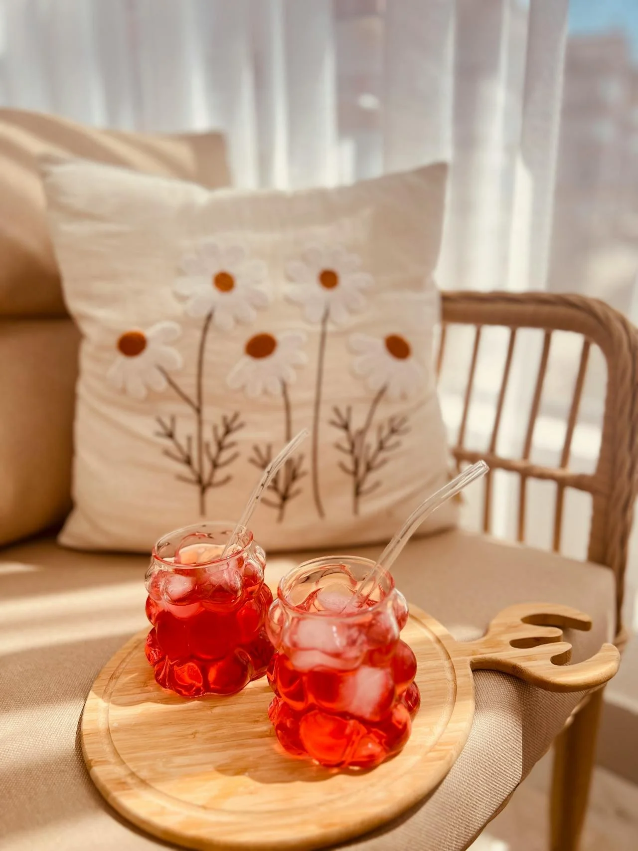 Two glasses of red-colored beverage with ice and straws on a wooden tray, with a decorative pillow featuring daisy flowers on a sofa in the background.
