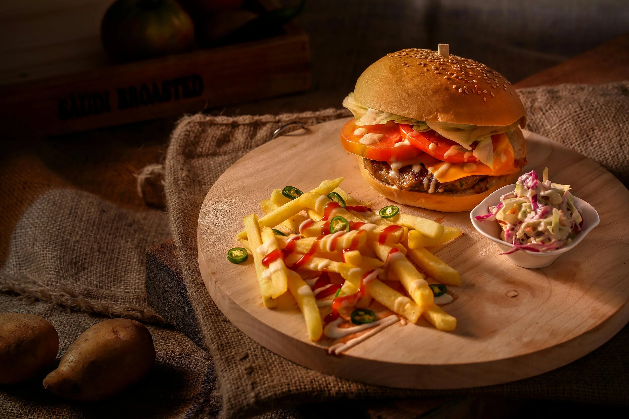 A cheeseburger with lettuce, tomato, cheese, and a beef patty in a sesame seed bun, served with French fries topped with ketchup and sliced jalapeños, accompanied by a side of coleslaw on a round wooden cutting board.