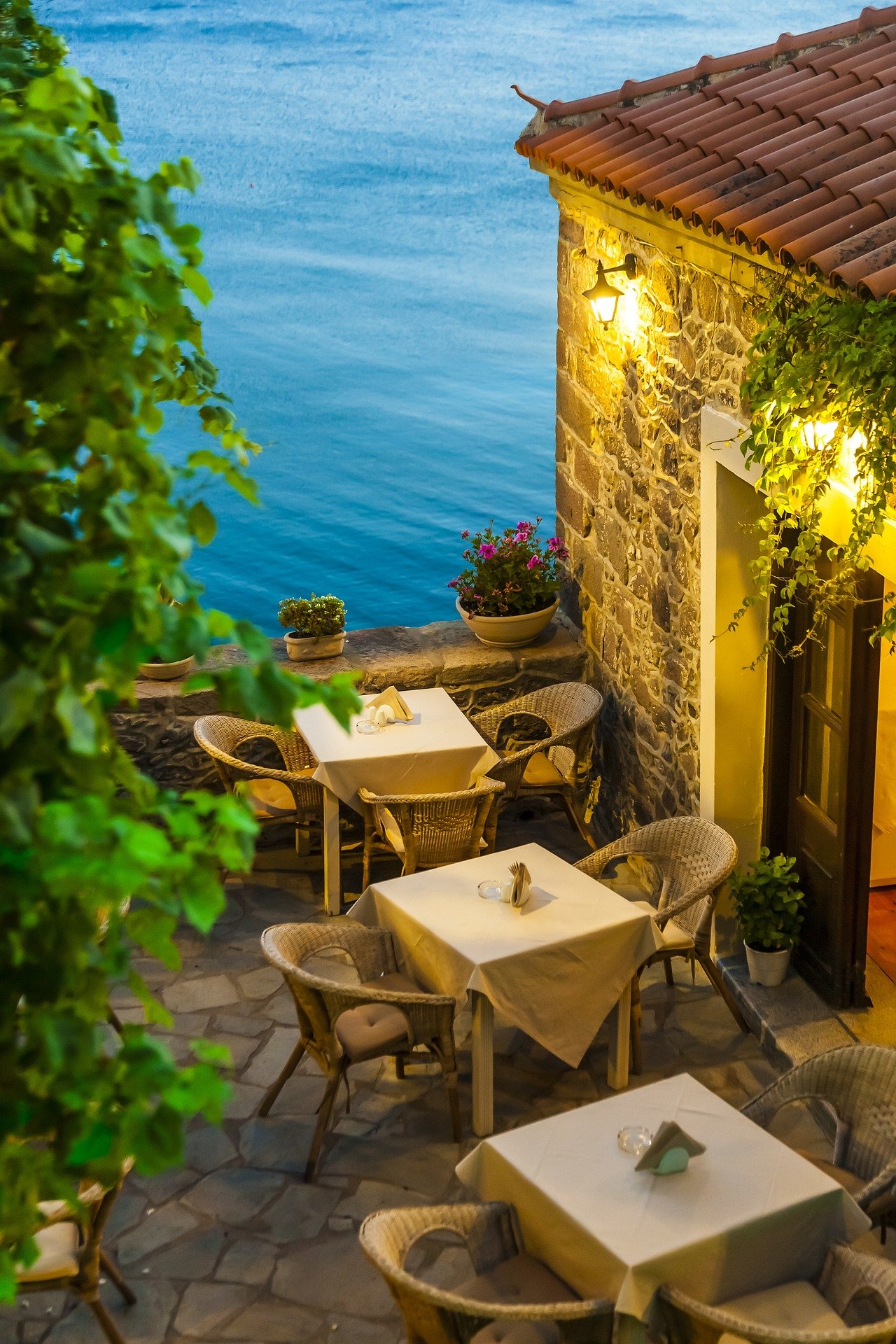 Outdoor dining area with four tables covered with cream tablecloths, surrounded by wicker chairs, overlooking a body of water at twilight. There are potted plants and flowers on the stone wall, warm yellow lighting, and a rustic stone building with a