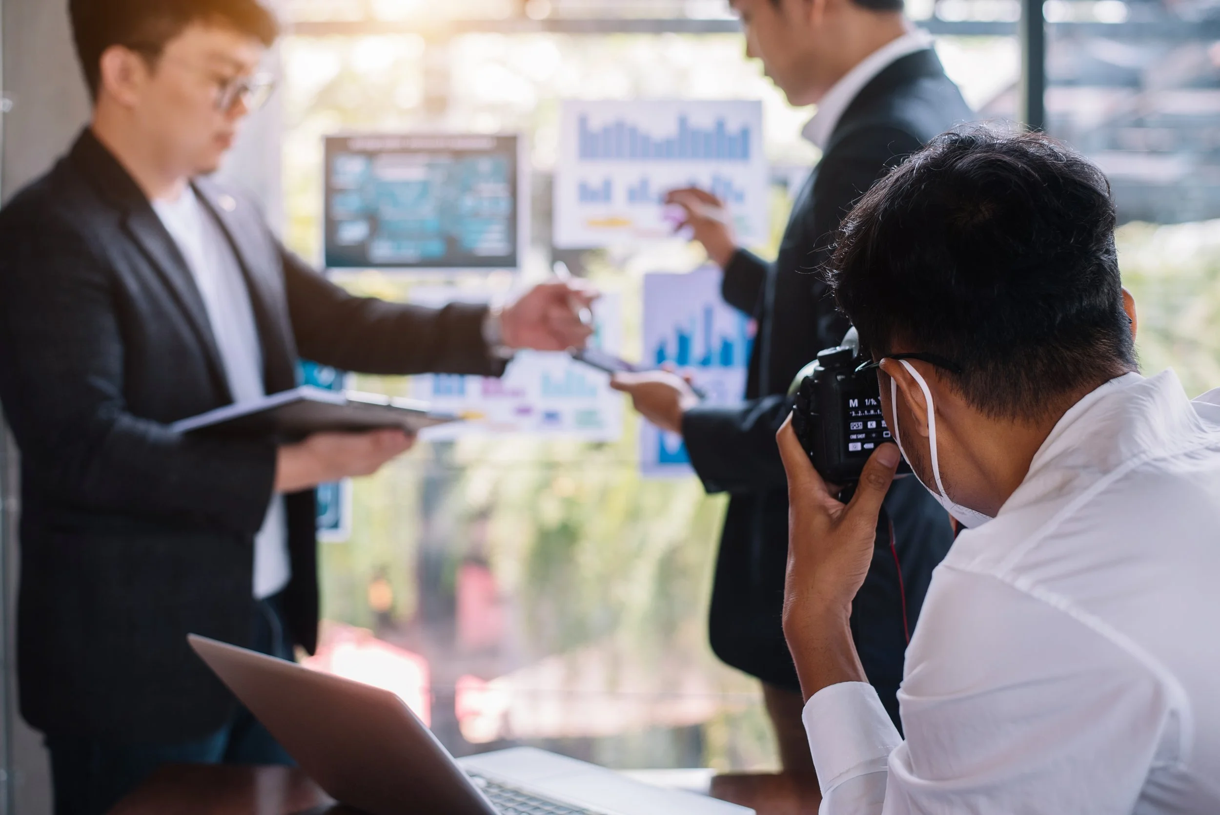 A group of business professionals, including a photographer, during a presentation or meeting with charts and graphs displayed on screens and posters in the background.