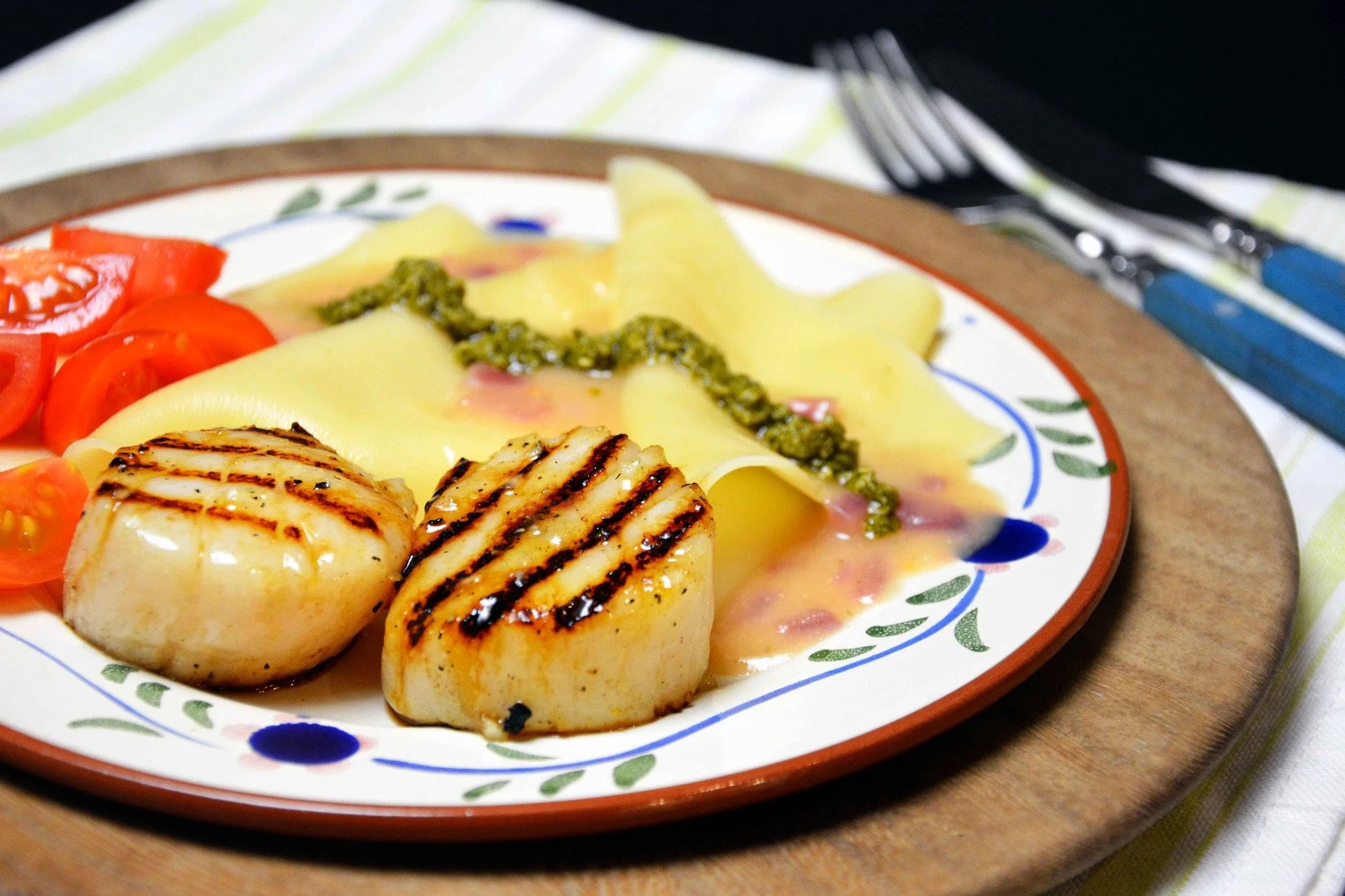 Close-up of grilled scallops with grill marks, served on a plate with cherry tomatoes, pasta, and green sauce, on a wooden tray with utensils in the background.