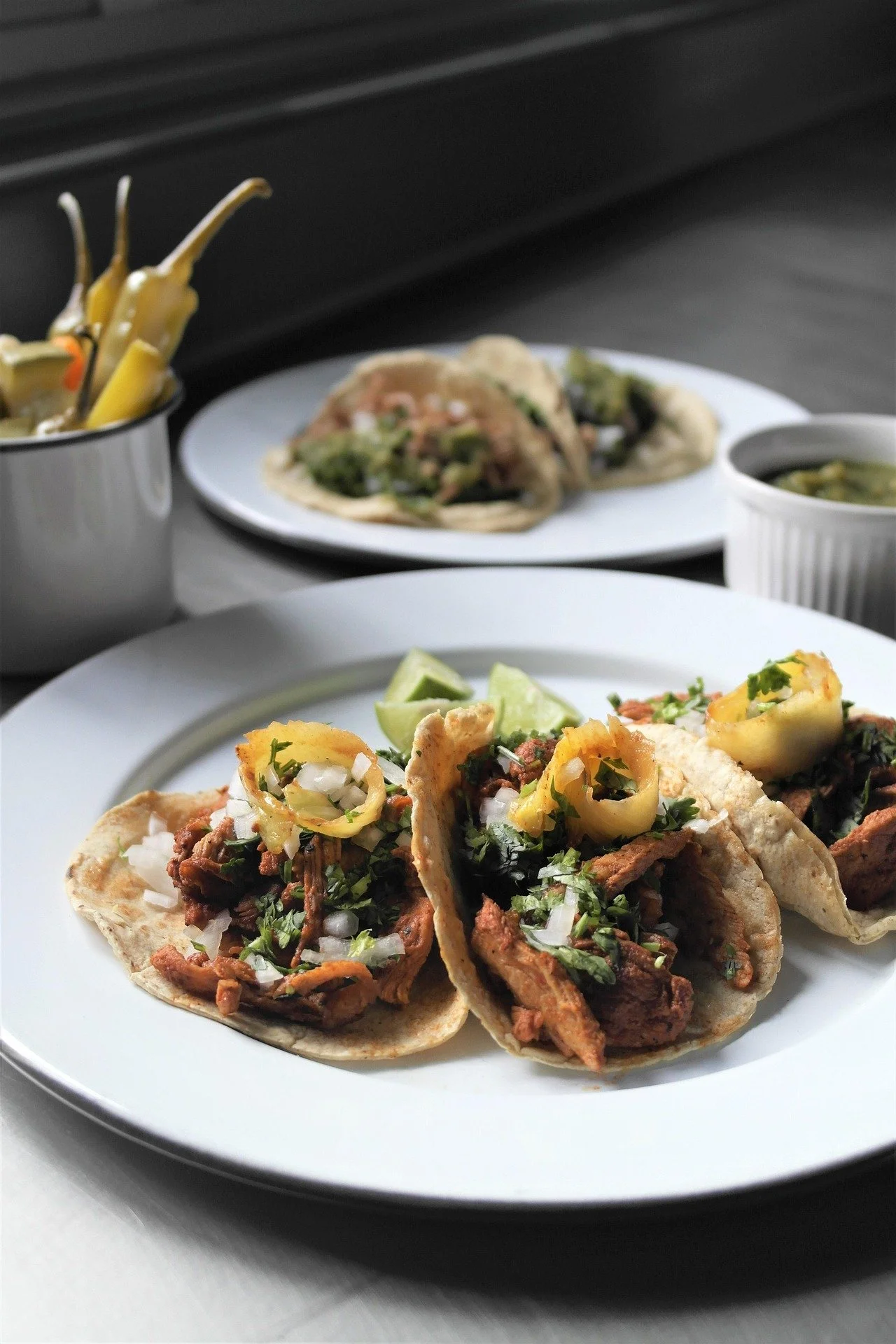 Close-up of three tacos filled with seasoned meat, chopped onions, cilantro, and topped with sliced jalapeños, served on a white plate with lime wedges. In the background, there are additional tacos, a small bowl of green sauce, a bowl of pickled veg