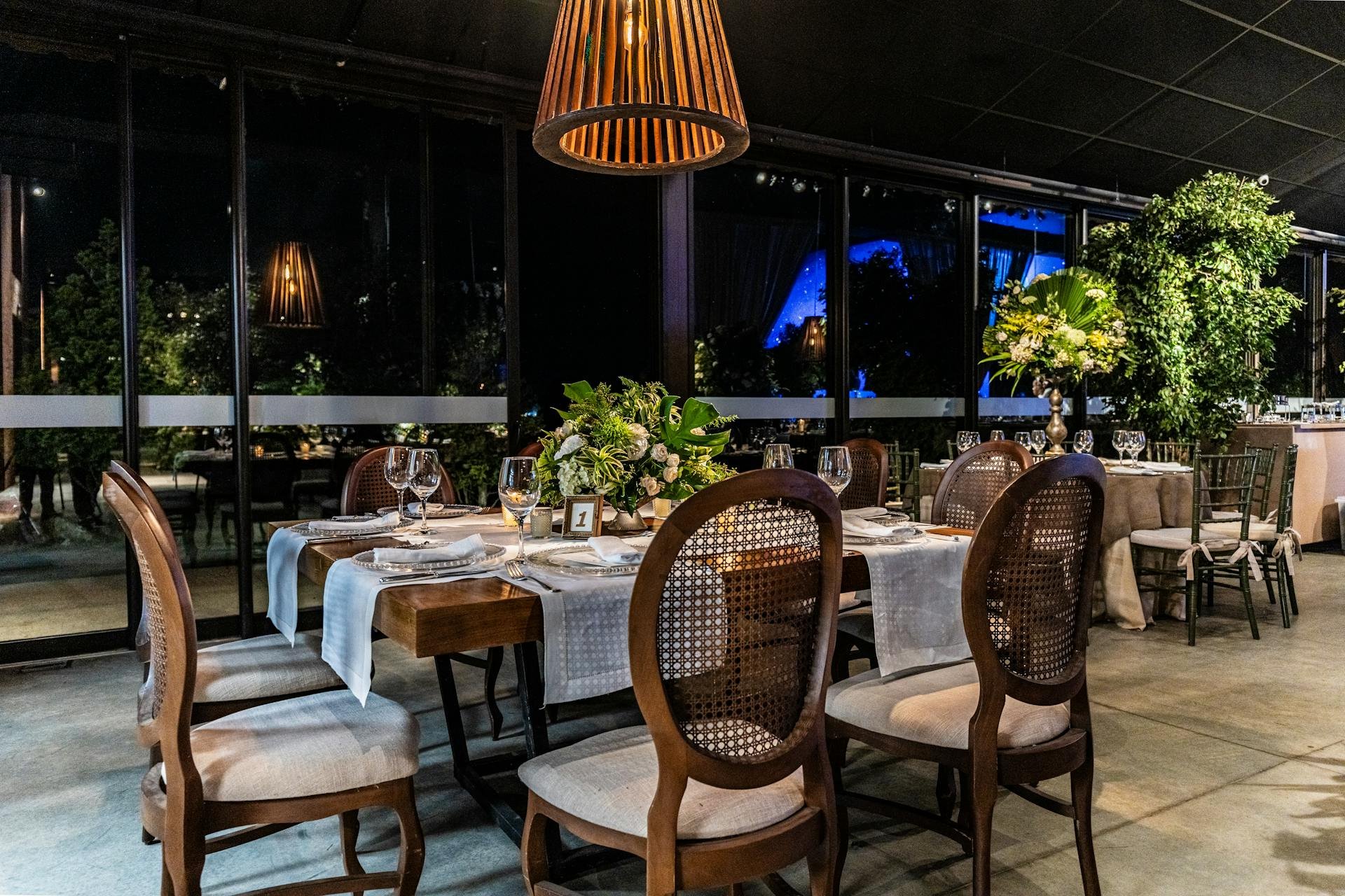 Elegant dining room set up with tables, chairs, and floral centerpieces in a glass-walled venue at night.