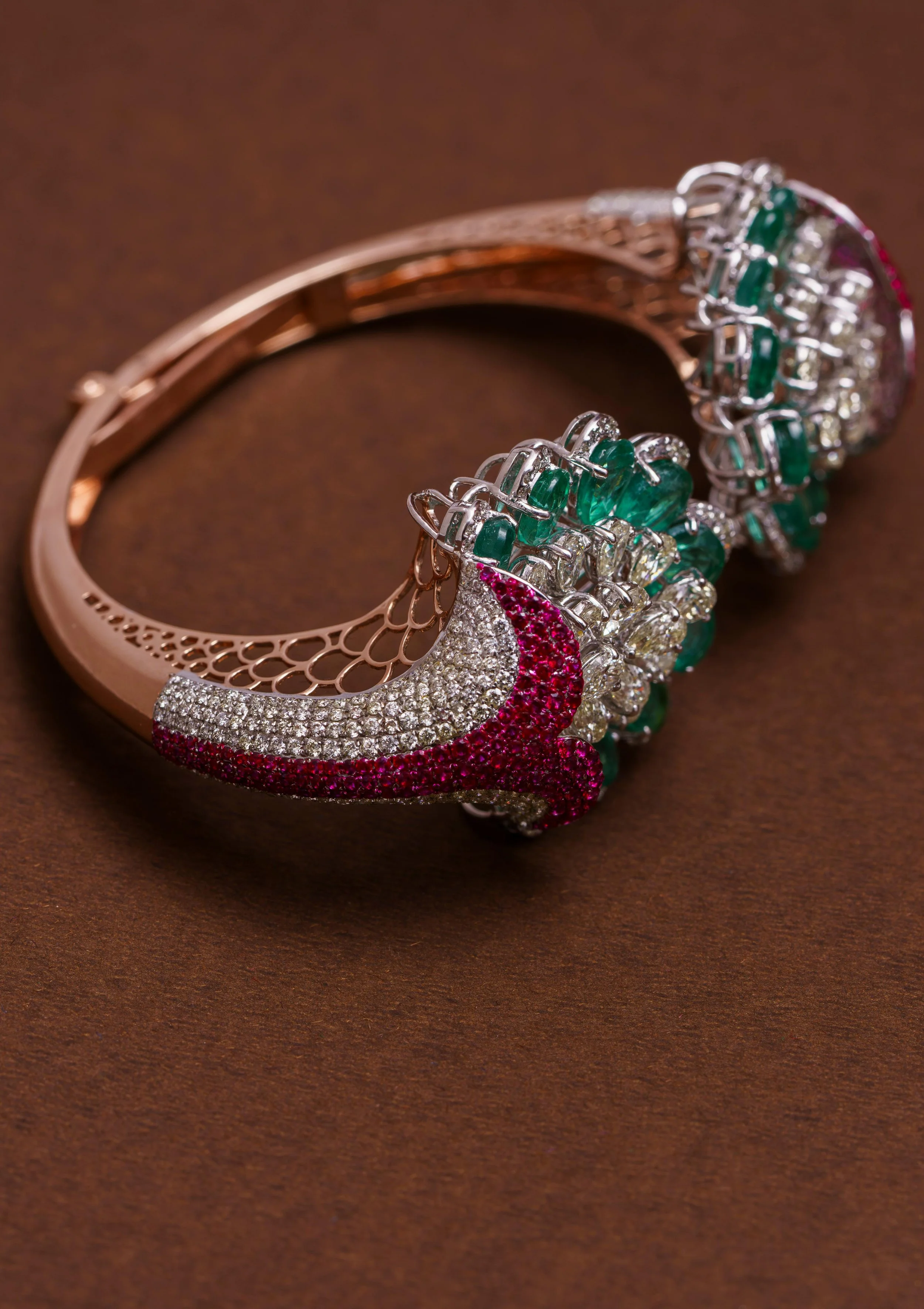 A decorative bracelet with a rose gold band, embedded with multiple small diamonds, featuring a layered, intricate design with red, white, and green gemstones.