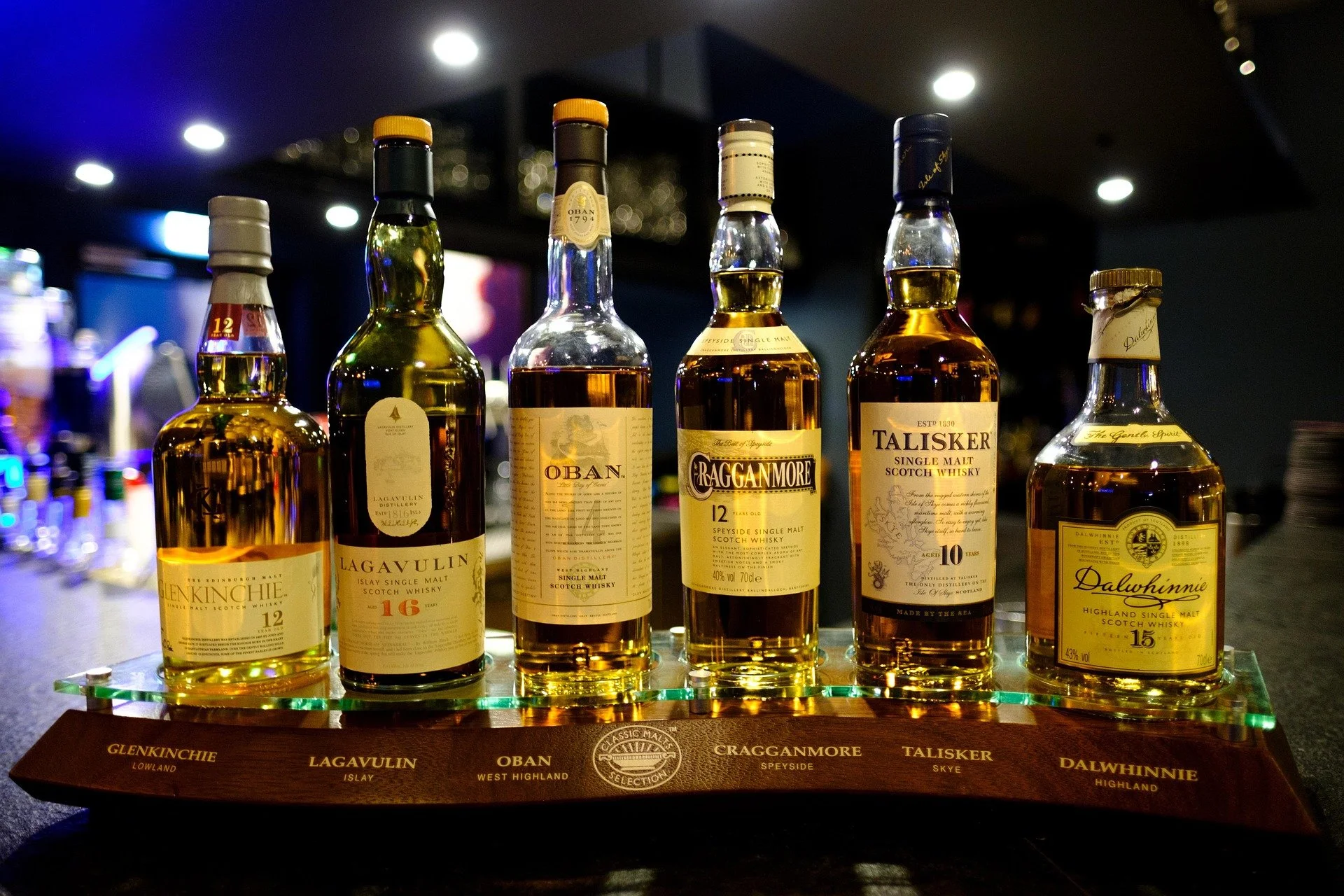 Six bottles of single malt Scotch whisky displayed on a high-backed wooden stand with brand names listed, including Glenkinchie, Lagavulin, Oban, Craggainmore, Talisker, and Dalwhinnie, in a dimly lit bar setting.