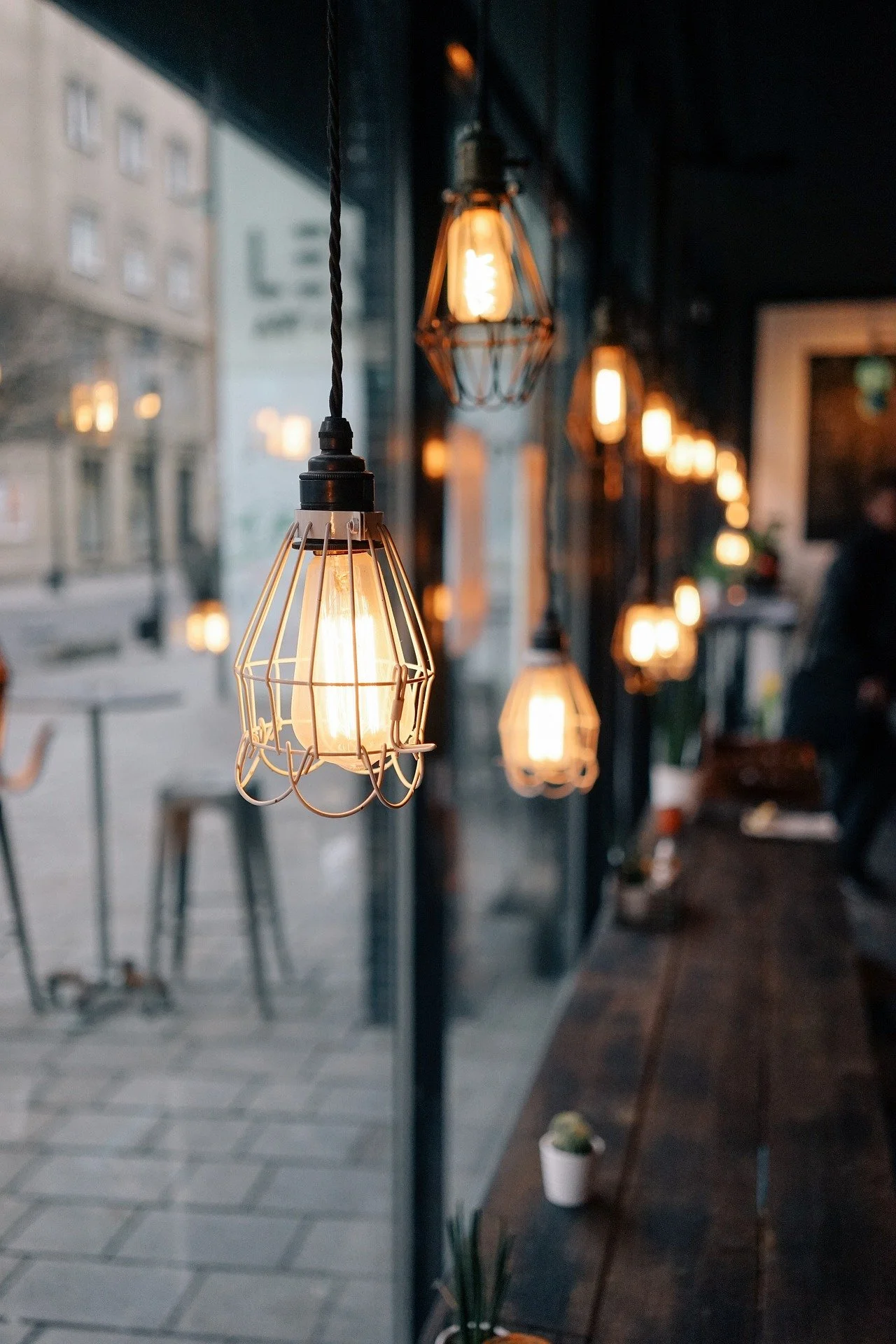 Indoor scene with hanging Edison-style filament light bulbs and a wooden table, with a view outside through large glass windows showing buildings and a sidewalk.