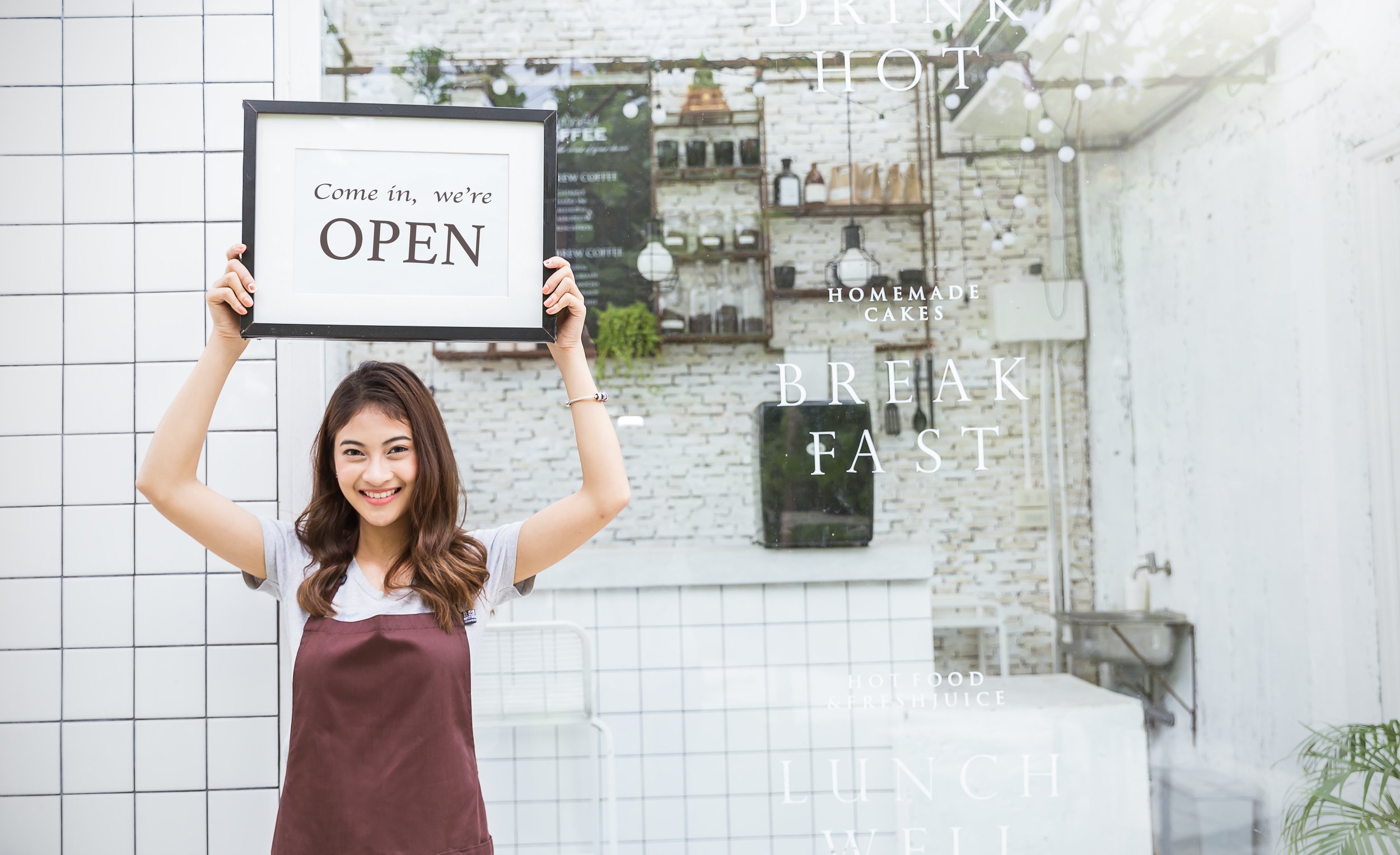 A young woman smiling and wearing an apron, holding a framed sign that says 'Come in, we're OPEN' in front of a cafe window with white text advertising homemade cakes, breakfast, lunch, and juice.