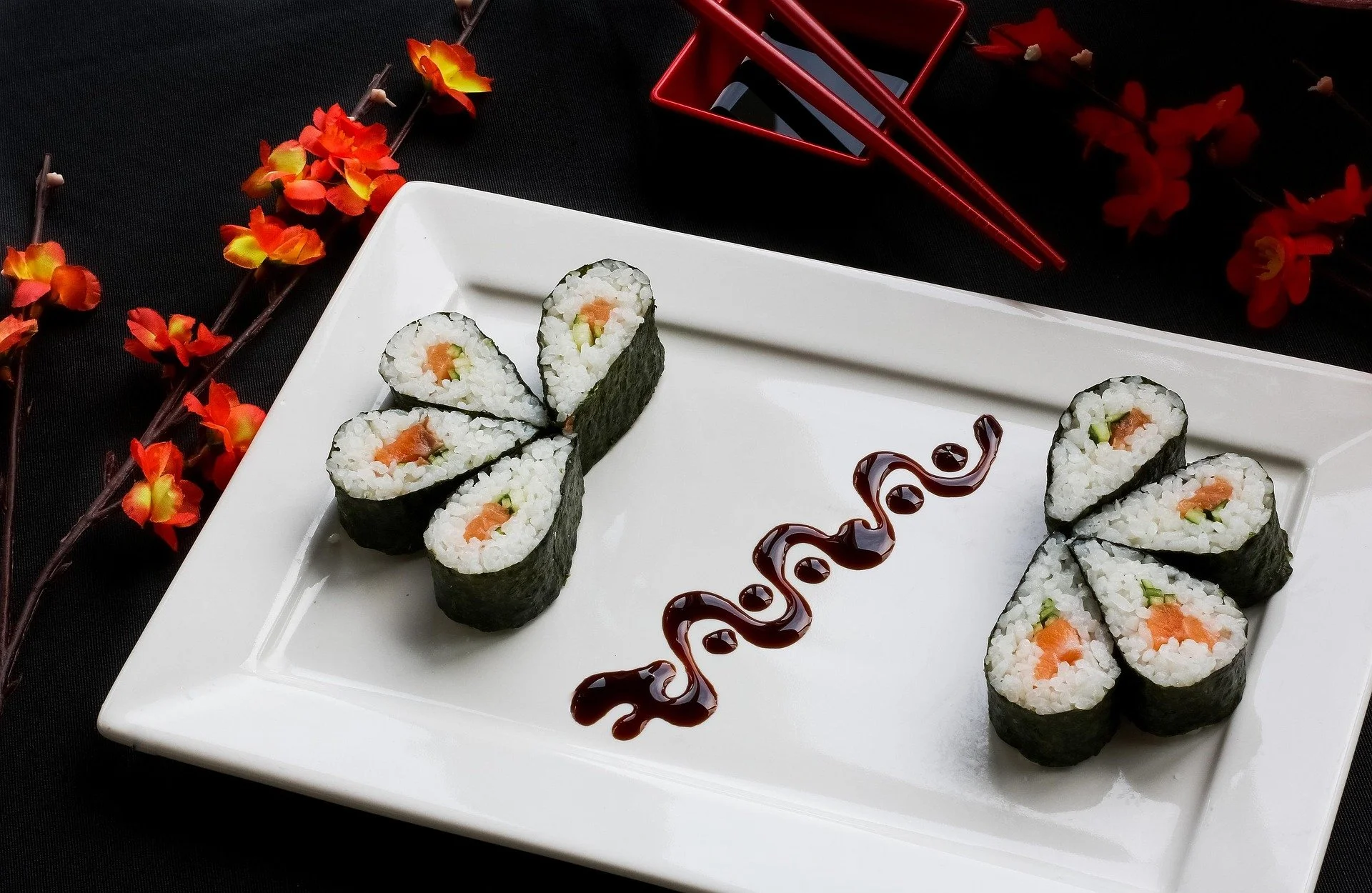 Plate of sushi rolls with soy sauce, red chopsticks, and decorative red and orange flowers on black background.