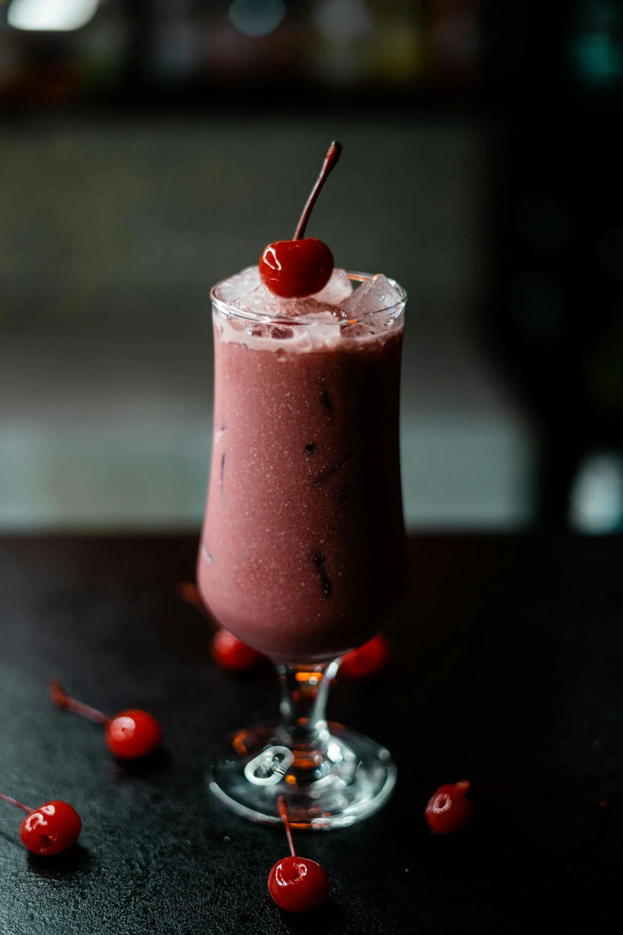 A pink cherry cocktail in a tall glass garnished with a cherry and surrounded by cherries on a dark surface.