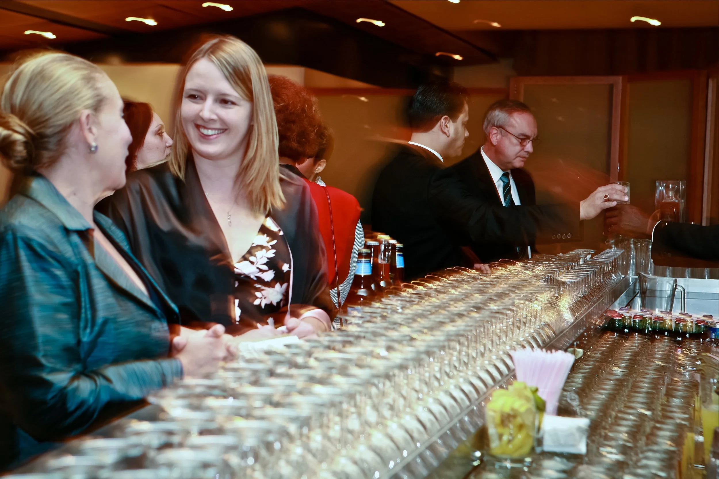 People standing at a bar with glasses and bottles, some pouring drinks, in a dimly lit setting.