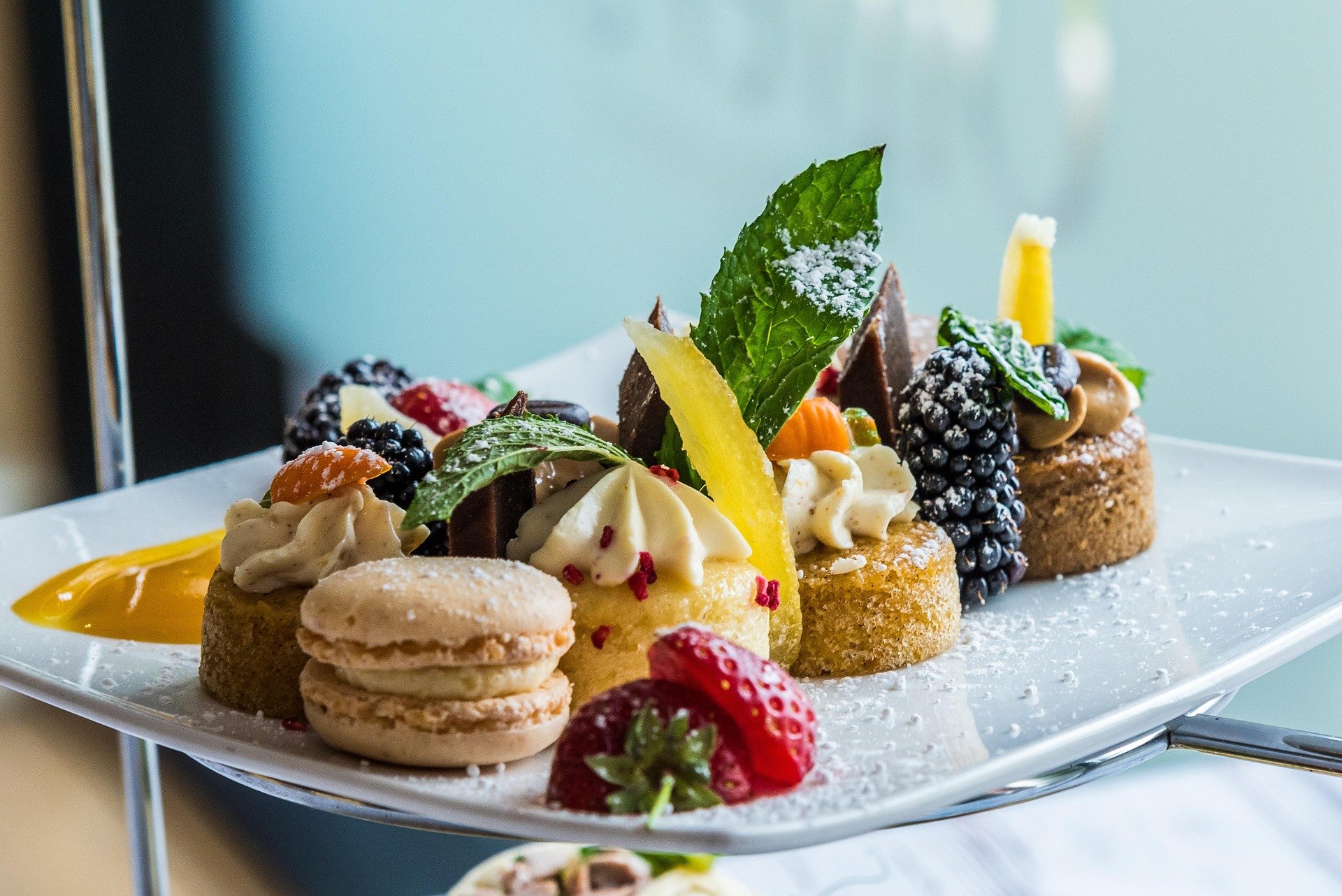 Assorted mini desserts on a white tiered platter, including macarons, cake squares, fresh berries, mint leaves, and decorative sauces.
