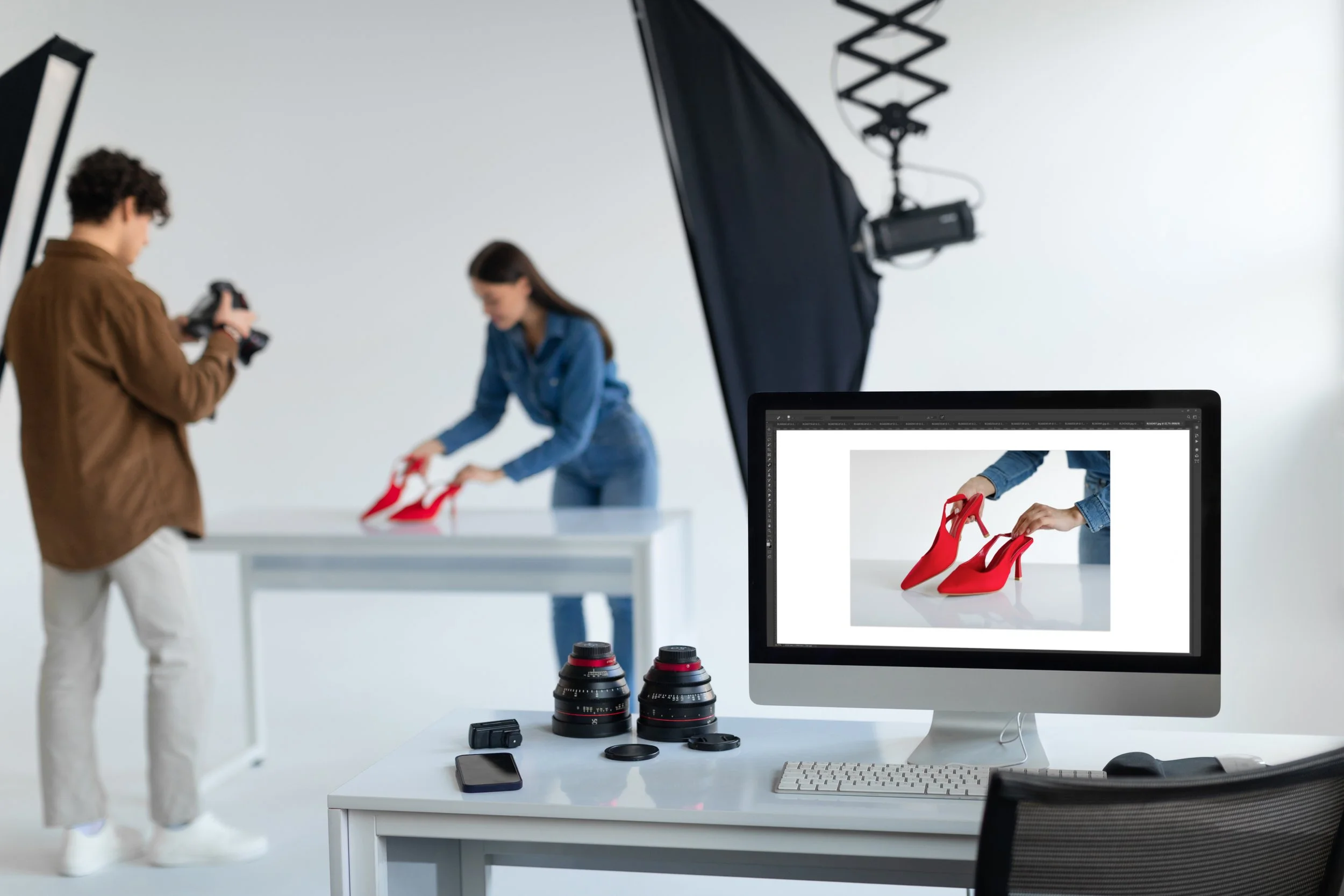 Photo of a photo shoot studio where a woman is displaying red high heels on a white table, with a photographer taking pictures and a computer displaying the same shoes. Equipment like lenses, smartphone, and lighting are visible.