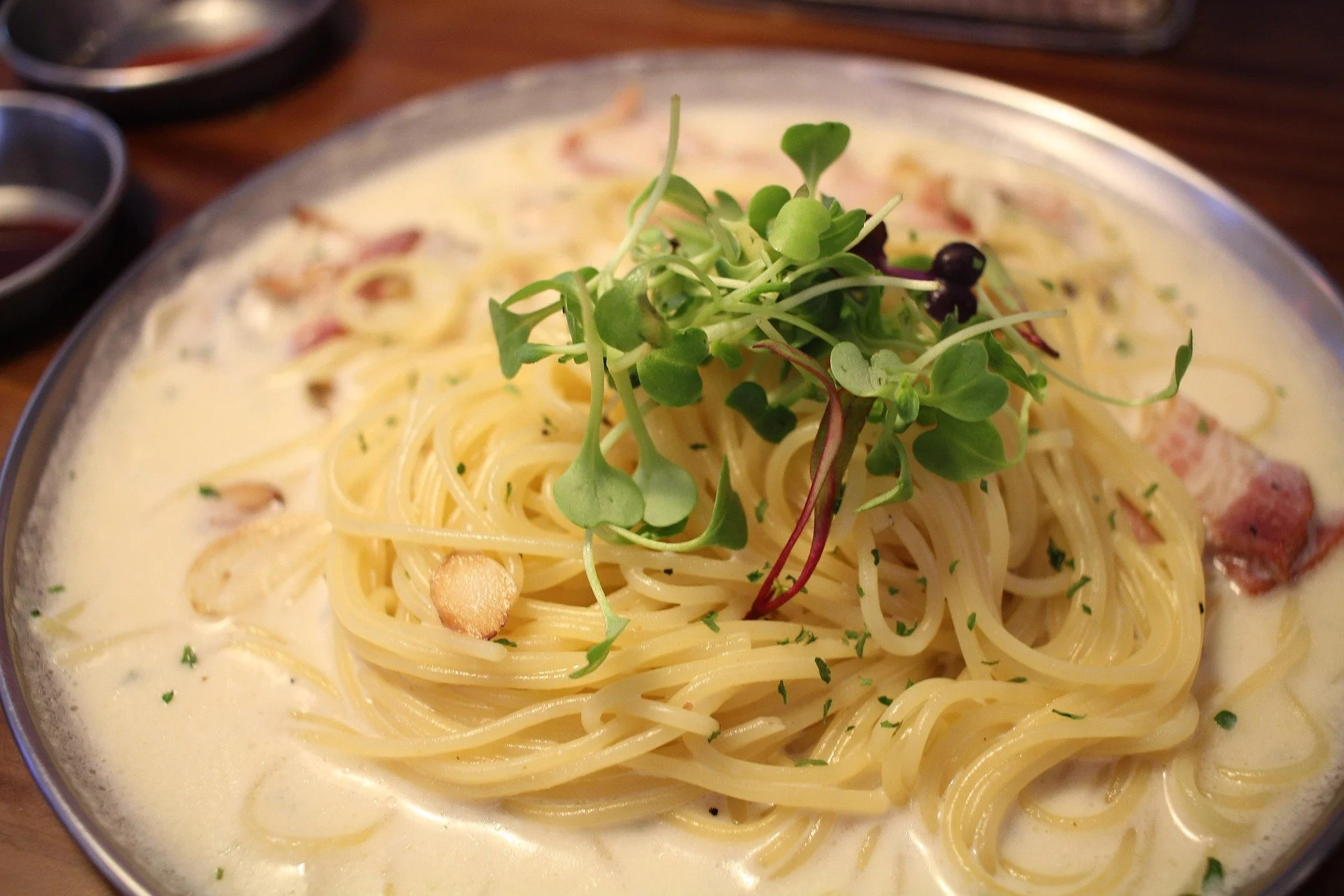 A plate of creamy spaghetti topped with microgreens and garnished with garlic slices. Some bacon strips are visible in the creamy sauce along with chopped herbs.