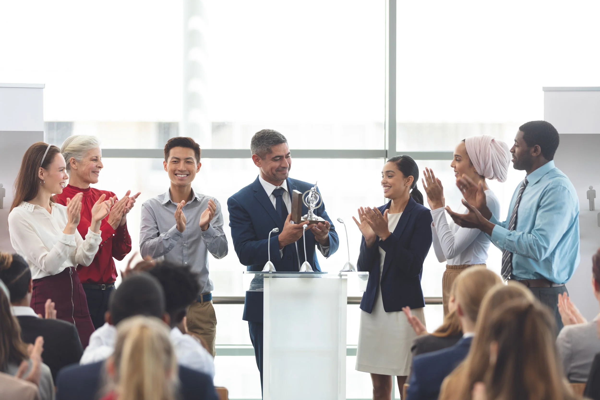 A diverse group of people at an awards ceremony, with a man in a blue suit receiving a trophy from a woman in a navy blazer, while others applaud and smile.