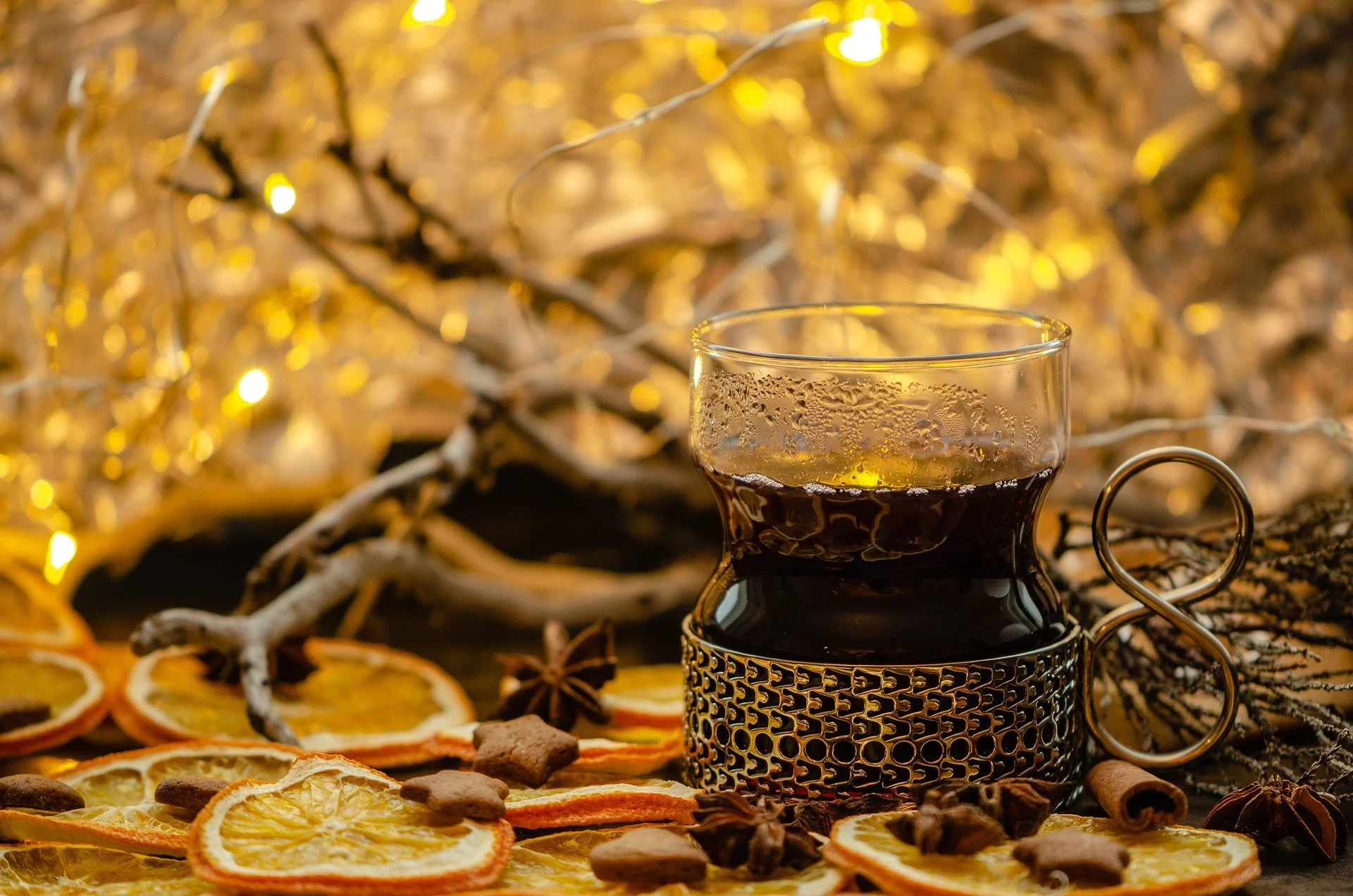 A glass of dark coffee with condensation, surrounded by dried orange slices, star anise, cinnamon sticks, and gingerbread cookies, with warm yellow string lights and twigs in the background.