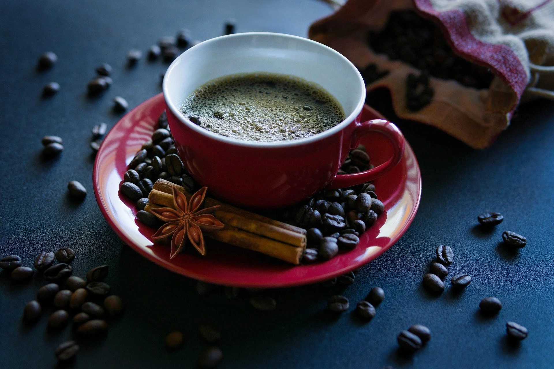 Cup of black coffee on red saucer with coffee beans, cinnamon stick, and star anise, with scattered coffee beans on dark surface.