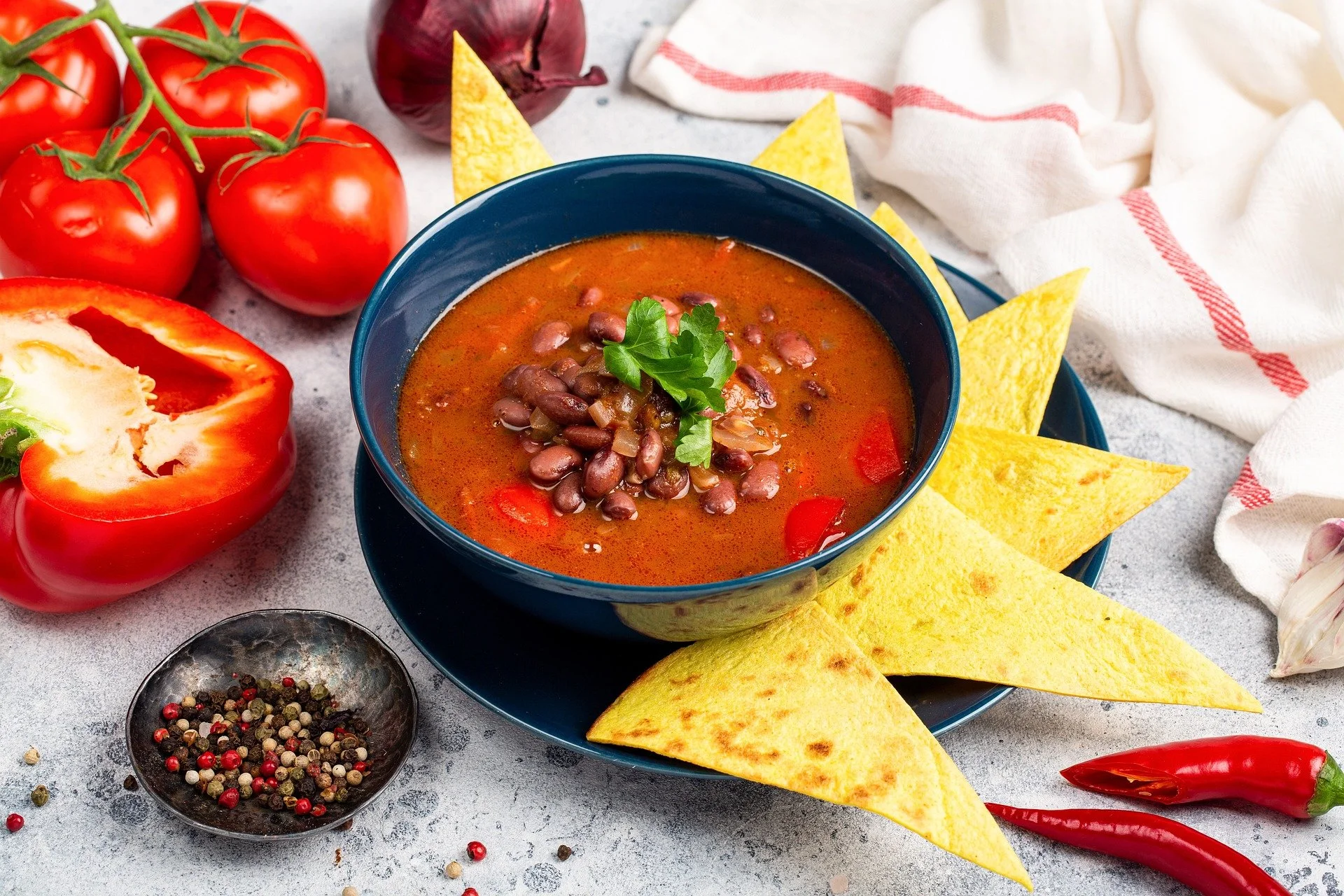 A bowl of Mexican black bean soup garnished with cilantro, surrounded by tortilla chips, red tomatoes on the vine, red chili peppers, a halved red bell pepper, a bowl of mixed peppercorns, a garlic bulb, and a white cloth with red stripes on a textur