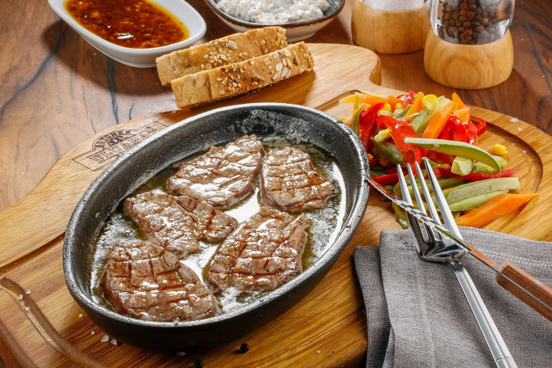 Grilled steaks frying in a cast iron skillet on a wooden cutting board, with a side of mixed vegetables including bell peppers, carrots, and celery, and a small dish of soy sauce or marinade in the background.