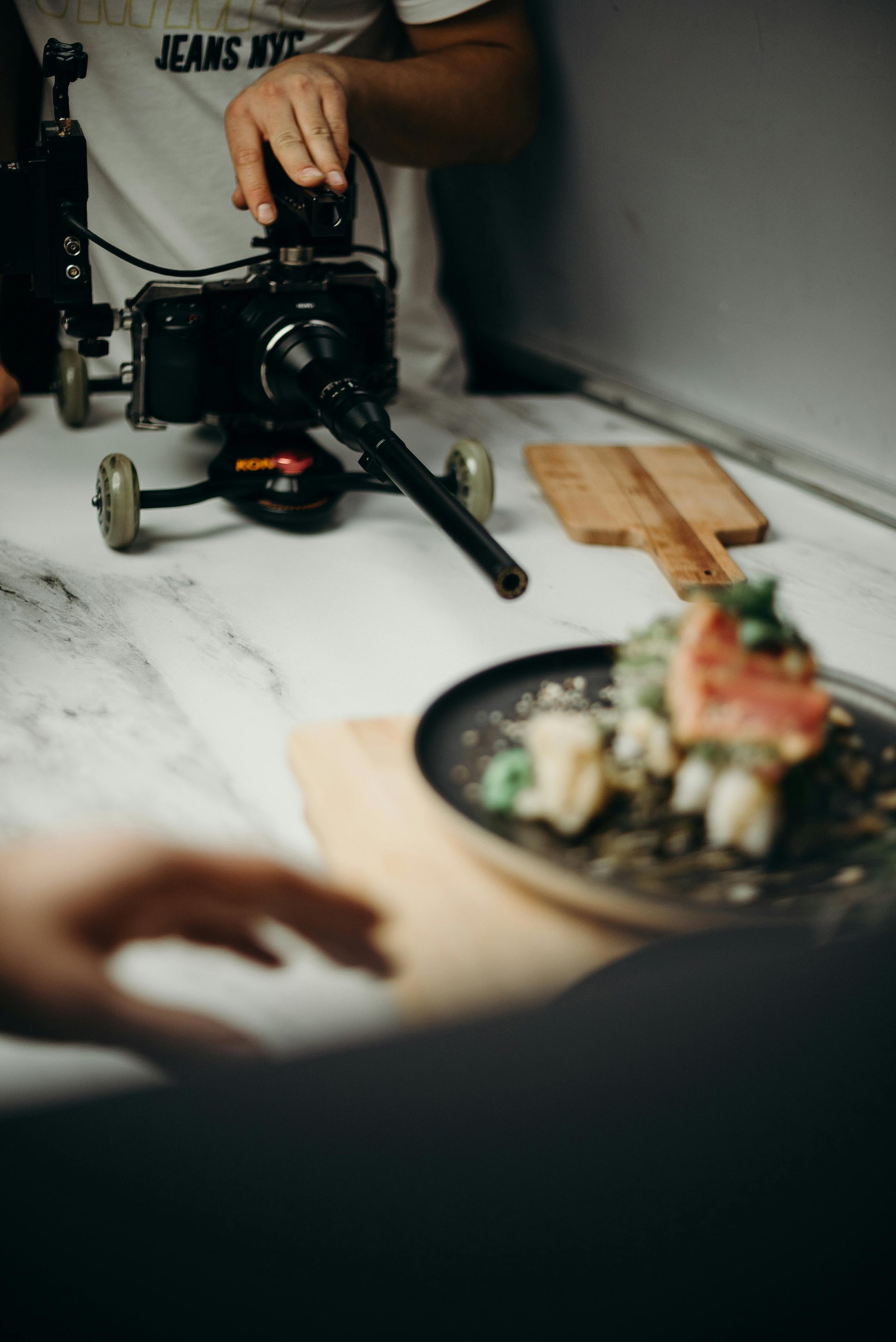 A photographer is using a camera mounted on a yellow skateboard to take pictures of a plated dish on a white marble countertop, with a wooden cutting board nearby.
