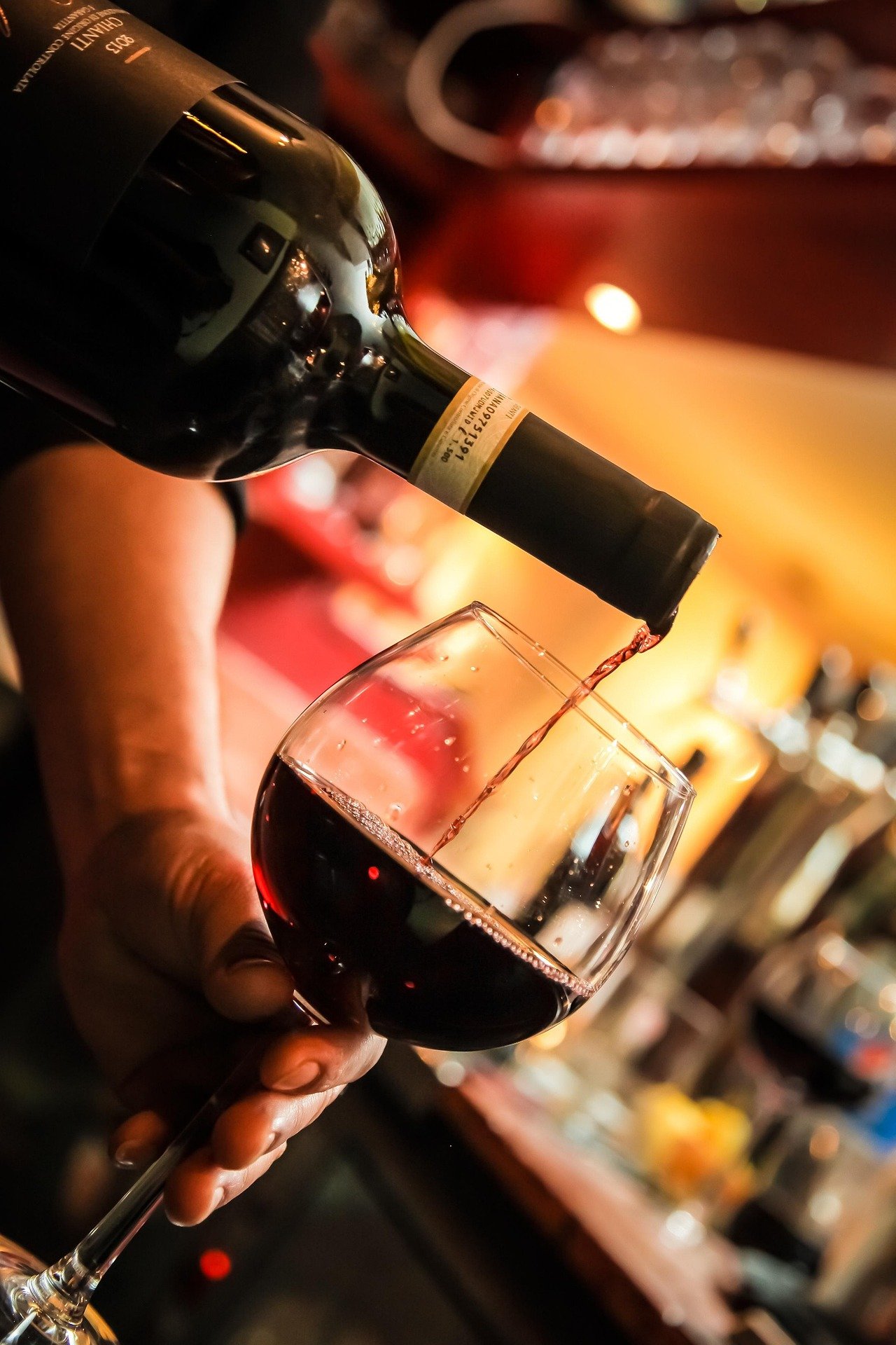 A person is pouring red wine from a bottle into a wine glass in a dimly lit setting.