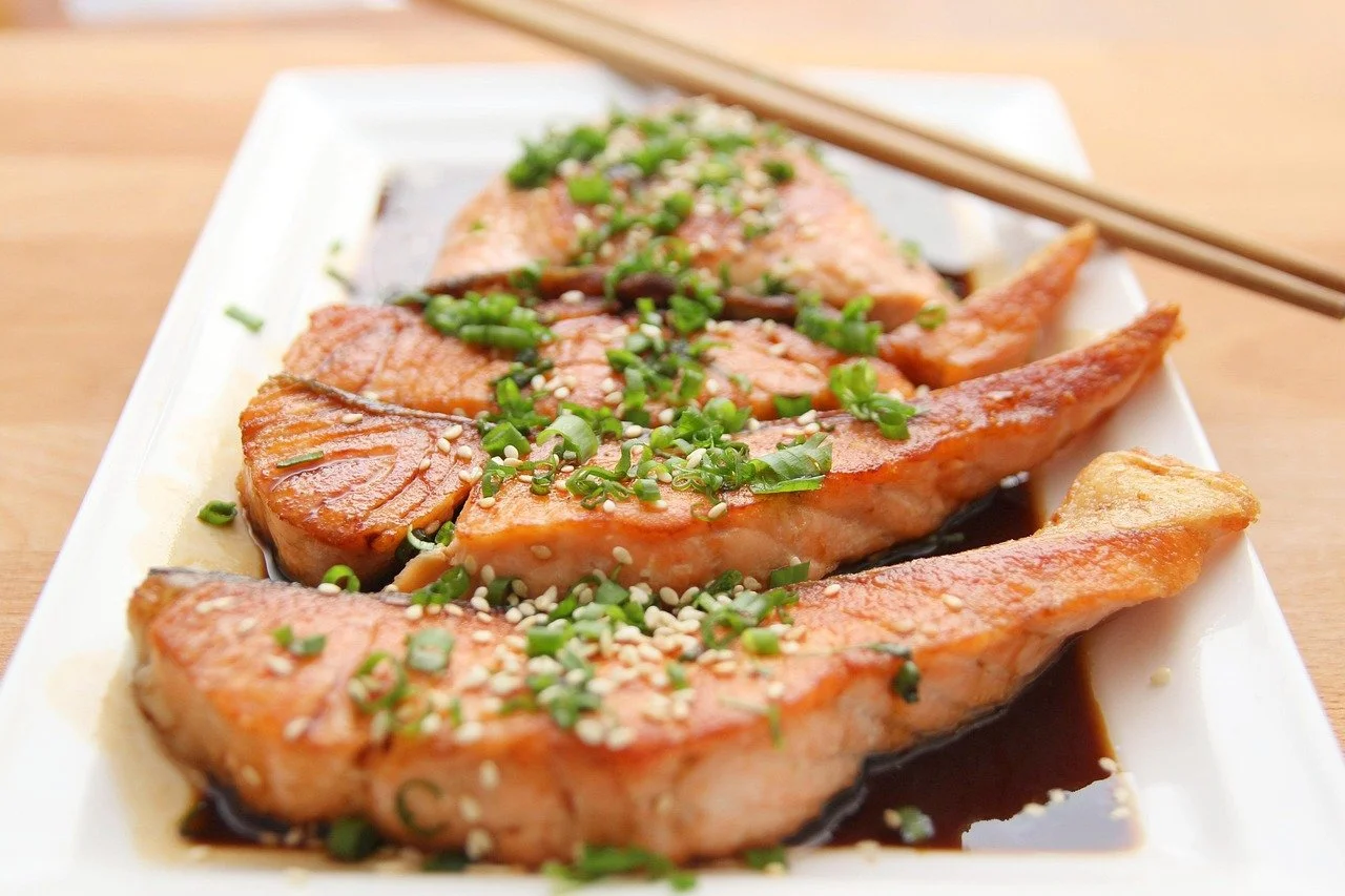 Close-up of grilled salmon fillets garnished with chopped green onions and sesame seeds on a white rectangular plate. Chopsticks rest on the side.